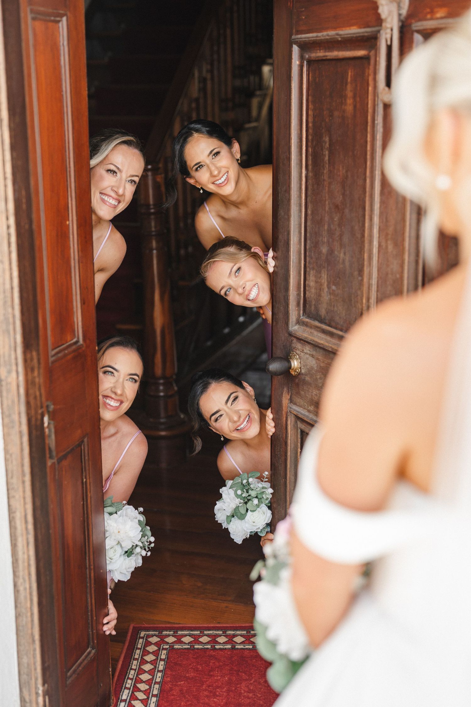 Bridesmaids peek playfully around wooden doorways in an elegant hallway with red carpet and ornate woodwork.