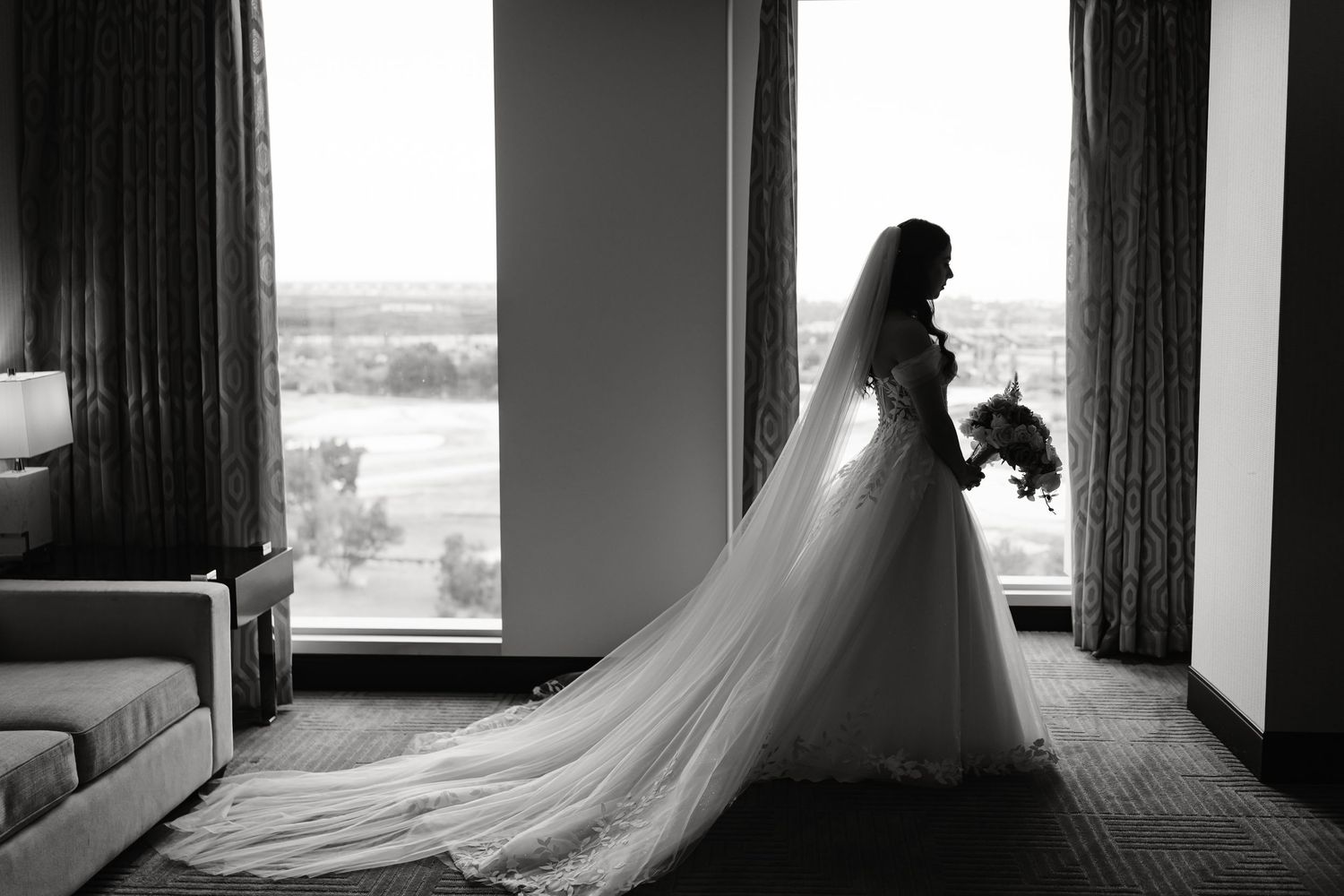 Black and white photo of a bride in a flowing gown and veil holding a bouquet while standing by large windows.