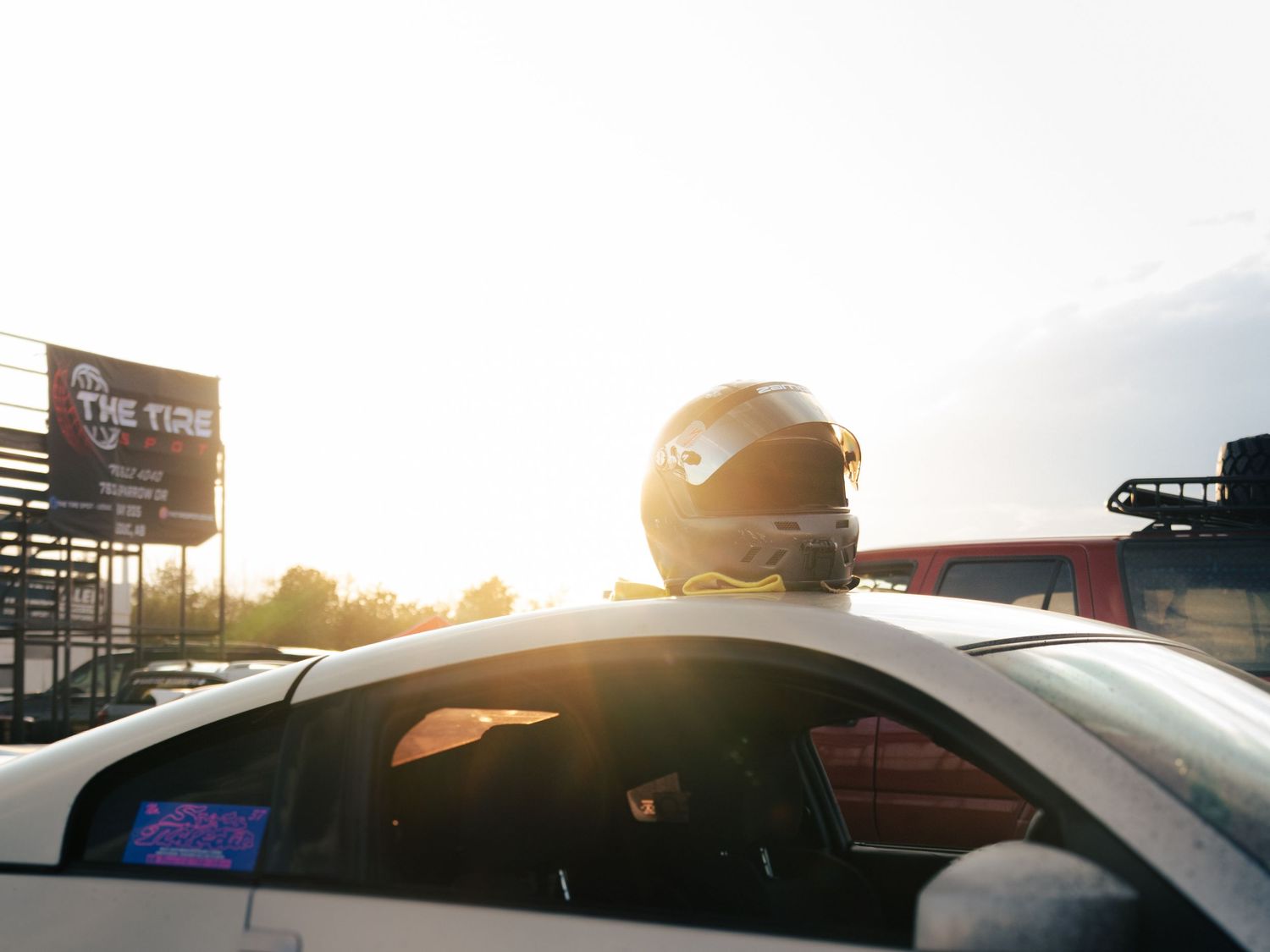 A race car driver wearing a helmet sits in a white car at a sunlit racetrack near The Tire sign.
