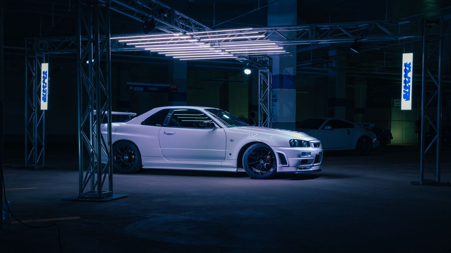 White Nissan Skyline R34 GT-R side profile under industrial lighting in a dark venue.