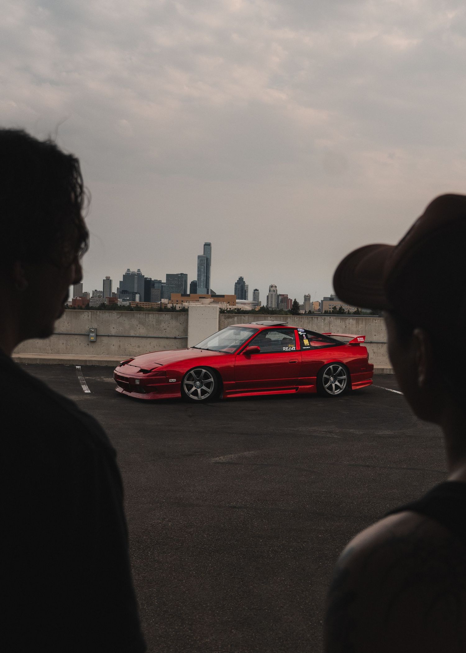 Two people watch a red Nissan 240SX on a rooftop parking lot with a city skyline in the background.