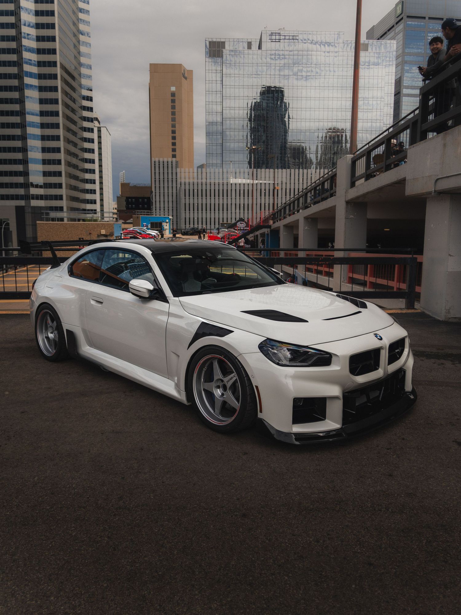 White modified BMW coupe with wide body kit and custom wheels parked in urban setting with city skyscrapers in background.