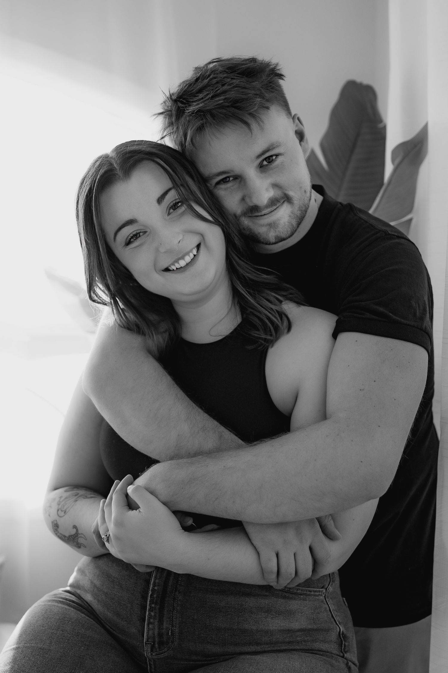Black and white portrait of a smiling couple embracing warmly indoors near a bright window.