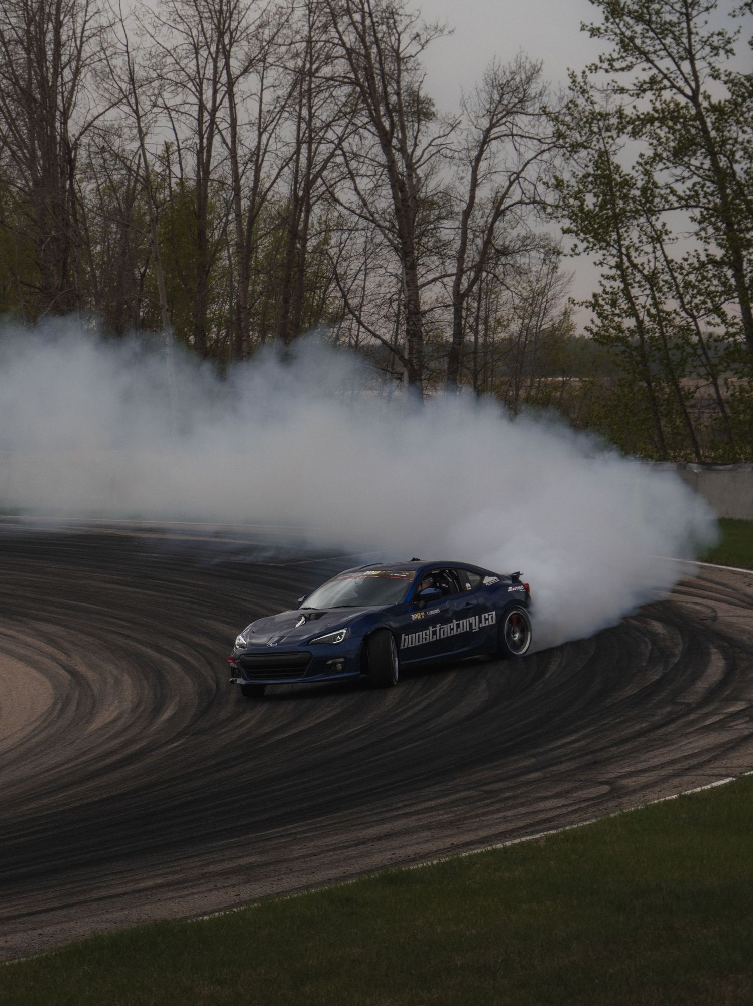 A blue sports car drifts around a track corner, producing large clouds of tire smoke, surrounded by bare trees.
