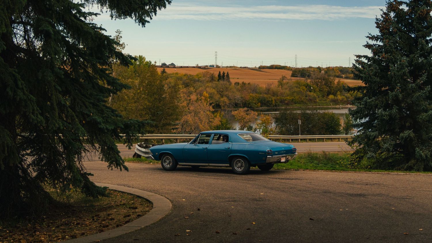 A classic blue muscle car is parked on a quiet road surrounded by trees with a scenic rural landscape in the background.
