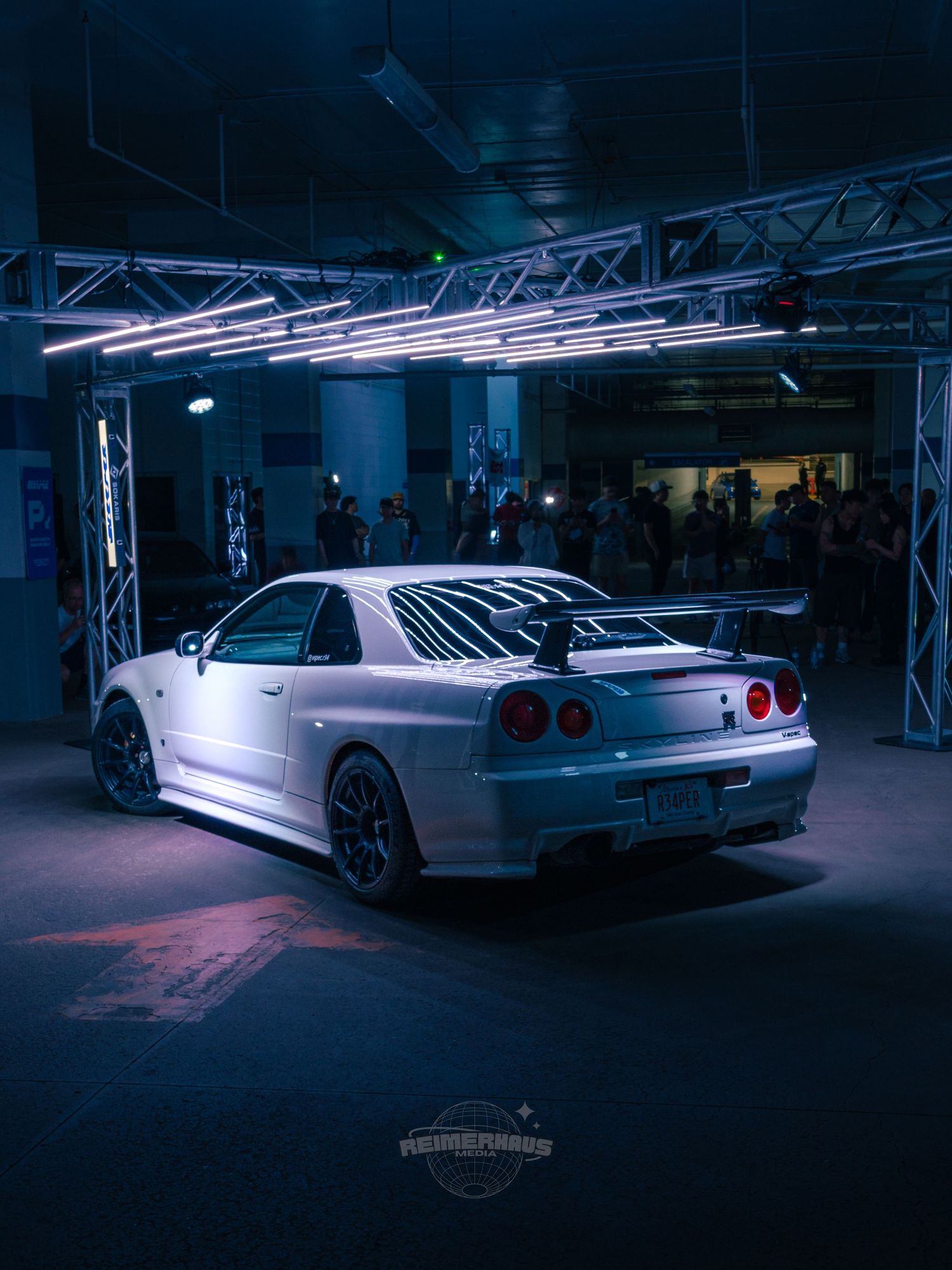 White Nissan Skyline R34 GT-R rear view under neon lights at an indoor car show.