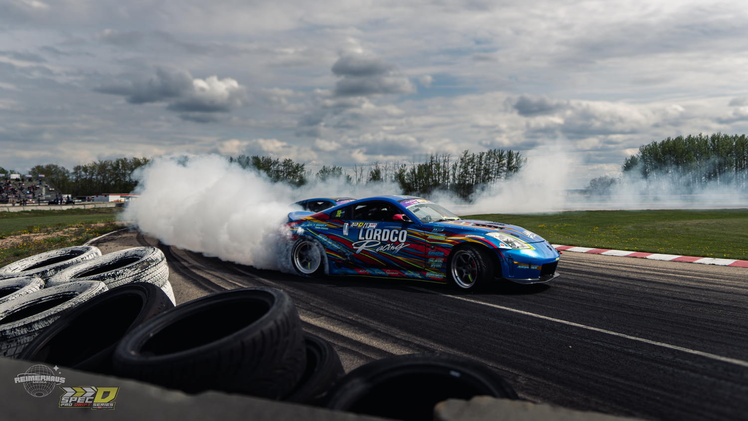 A race car drifts on a track, producing massive smoke clouds, with tires lining the course under a cloudy sky.
