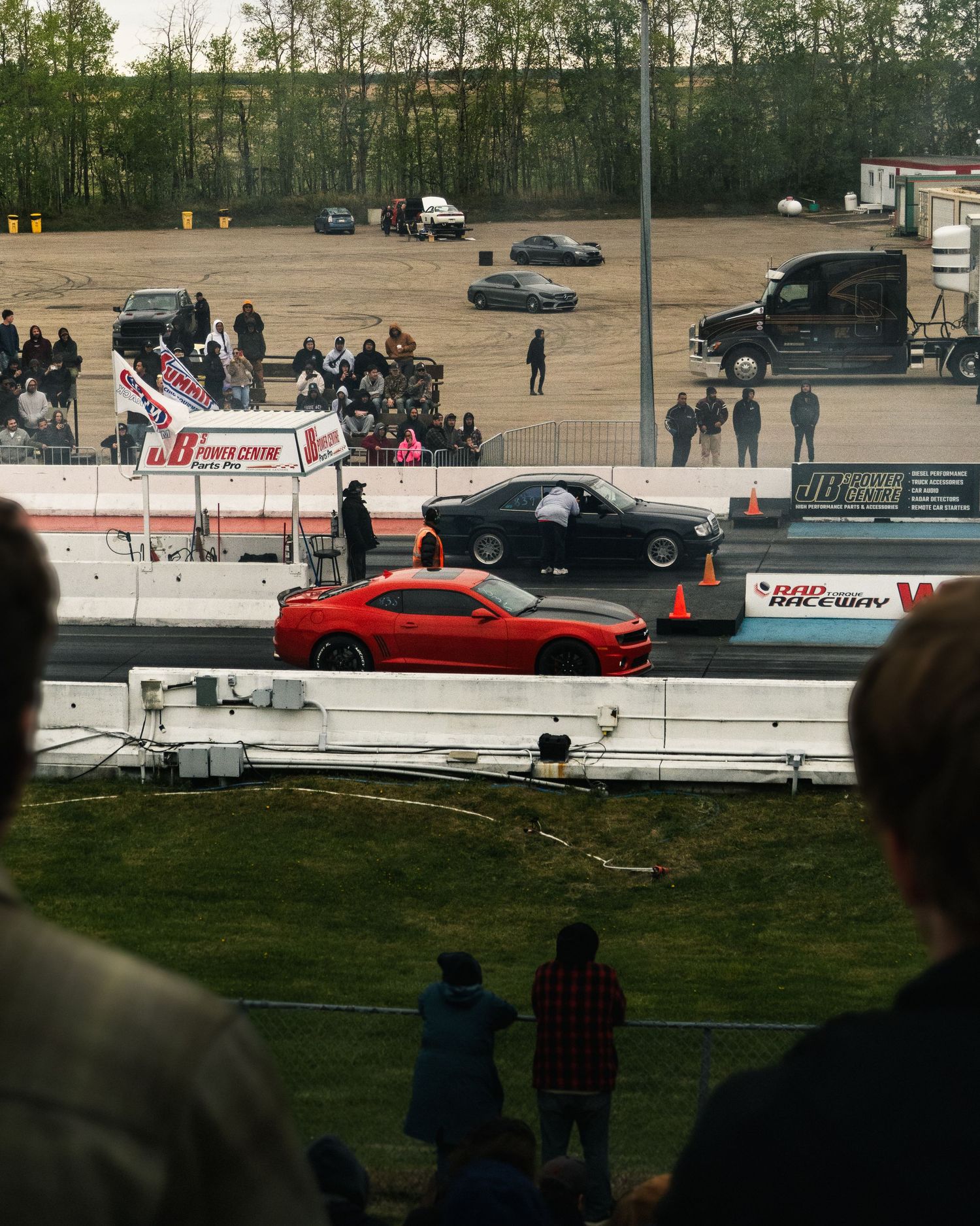 Two muscle cars drag racing on a track with spectators watching from the stands at an outdoor dragway.