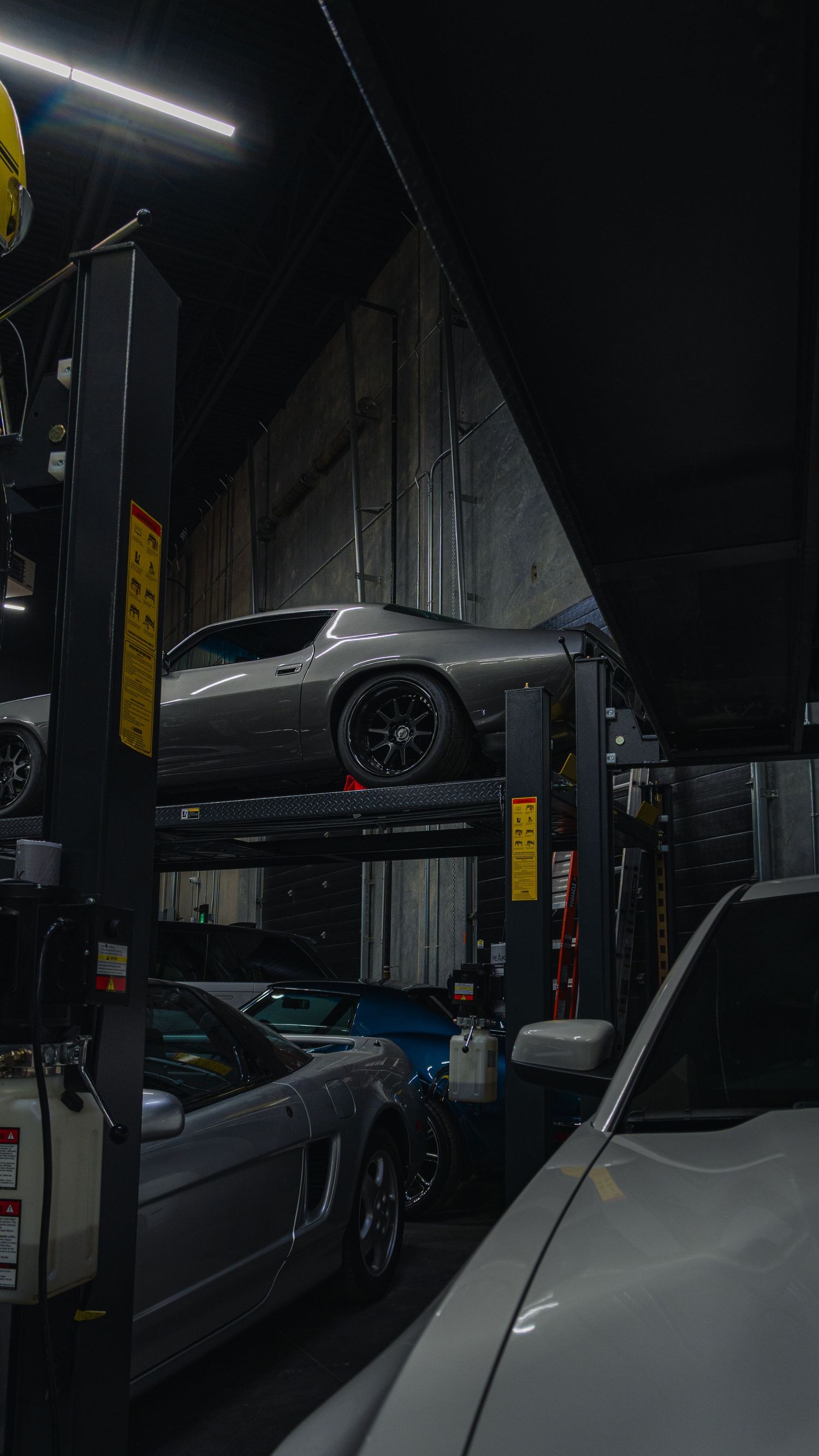 Dark auto repair shop with a silver muscle car elevated on a four-post lift above sports cars.