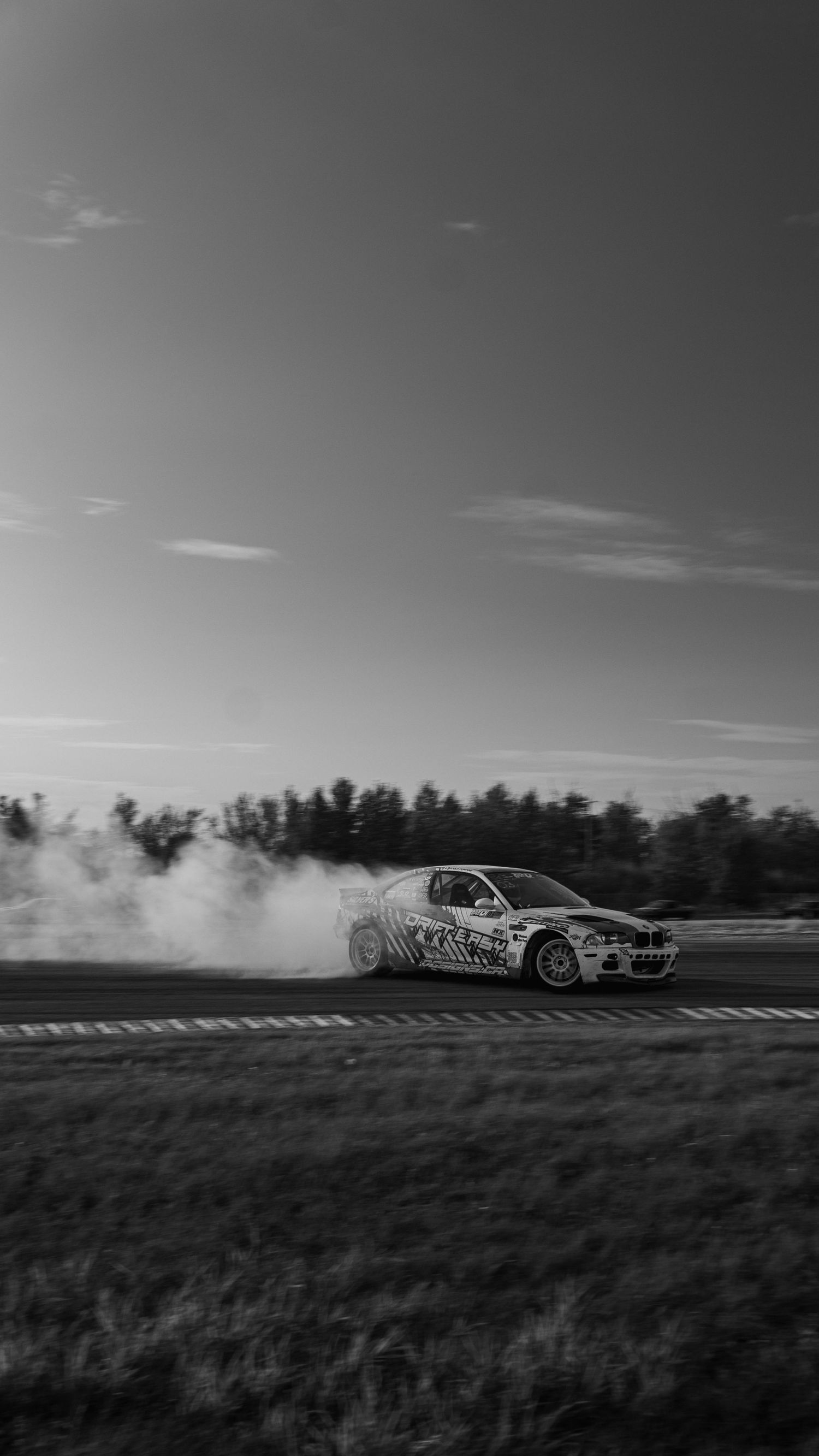 Black and white photo of a BMW drifting on a race track, leaving a trail of tire smoke against a dramatic sky.