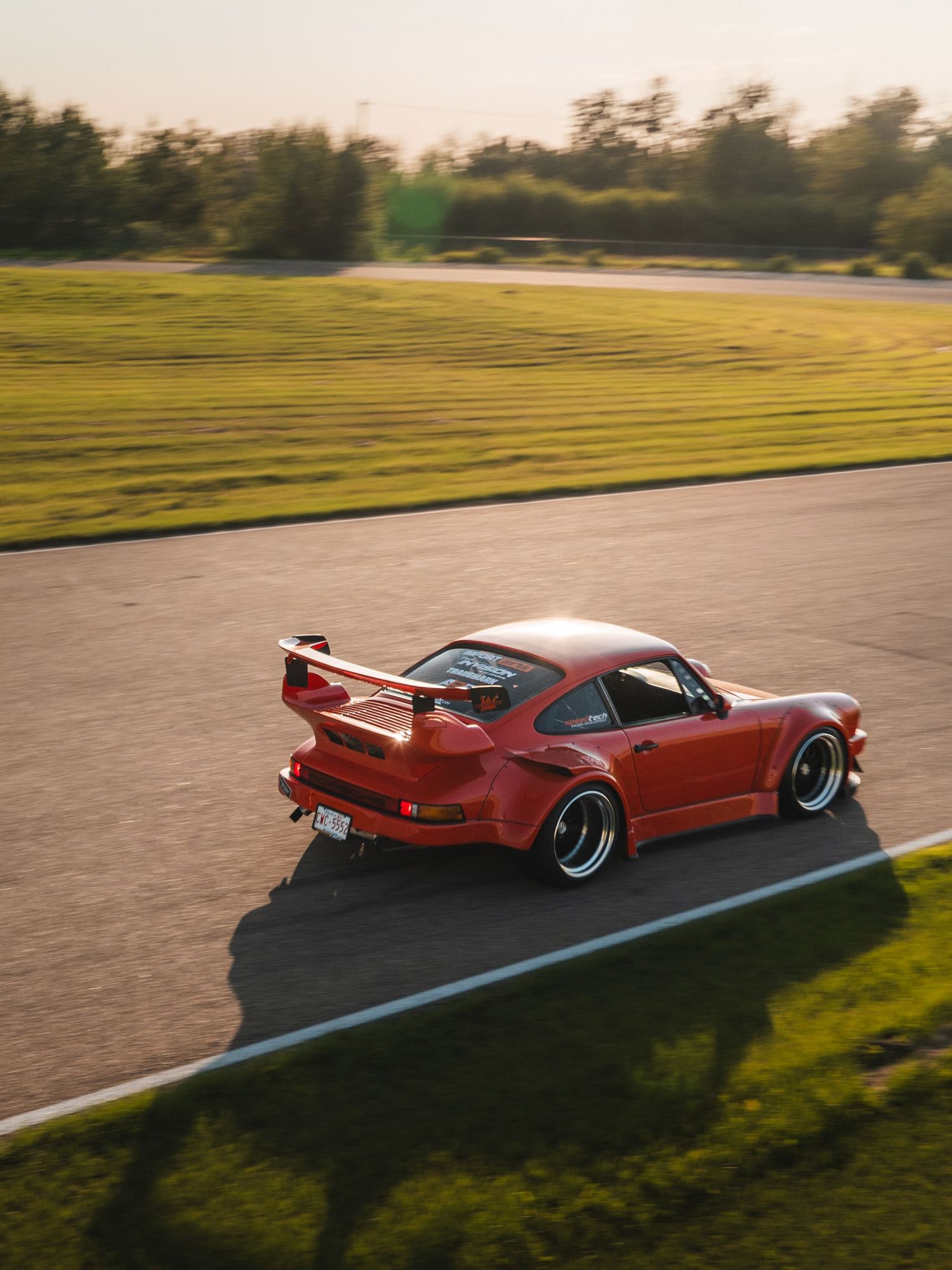 Red Porsche 911 RWB wide-body on track at golden hour, rear spoiler prominent, motion blur background.