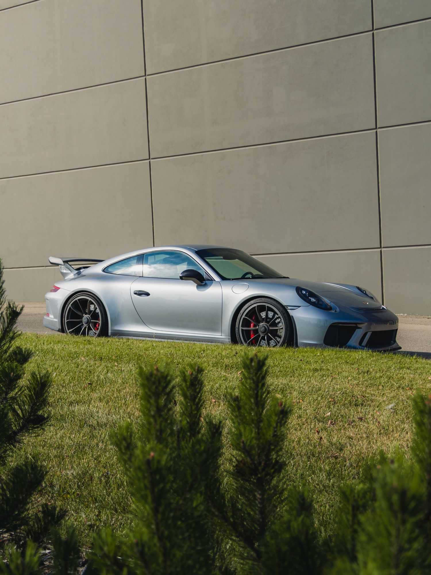 Silver Porsche 911 GT3 sports car parked beside a modern concrete wall with green grass and shrubs in the foreground.