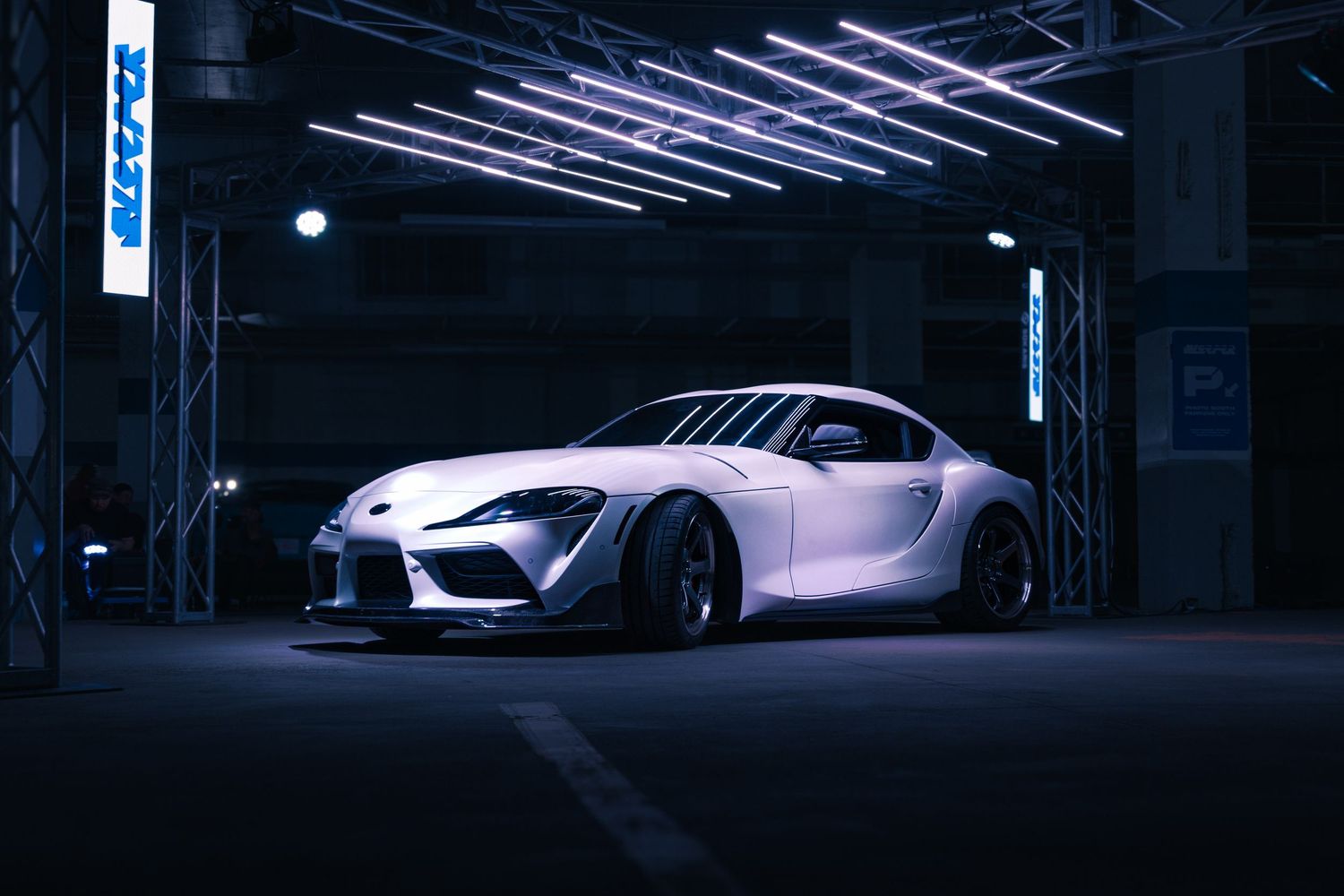 White Toyota GR Supra widebody kit parked in a dark garage under bright overhead lights.