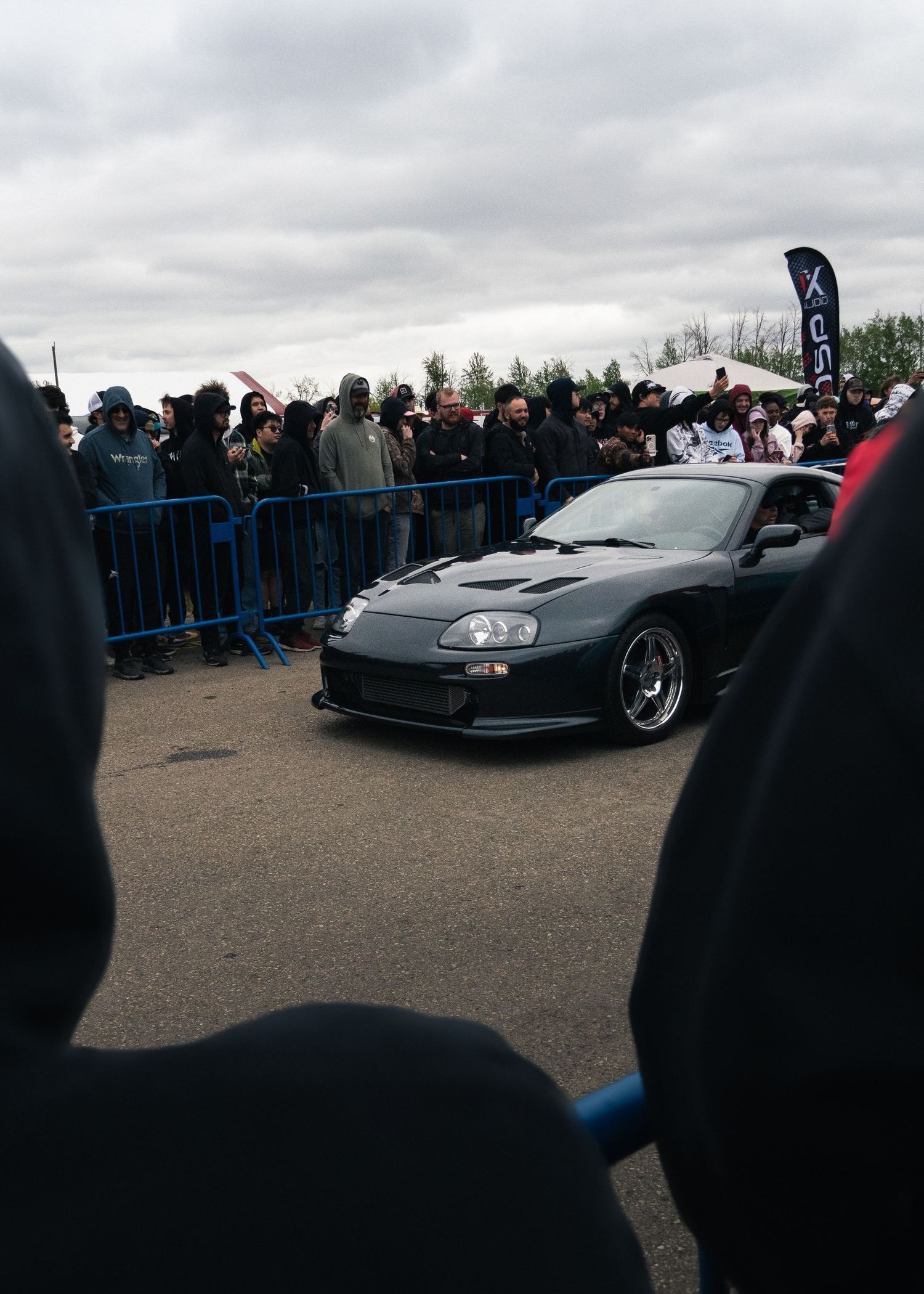 A dark gray Toyota Supra at a car show event with spectators behind blue barriers under an overcast sky.