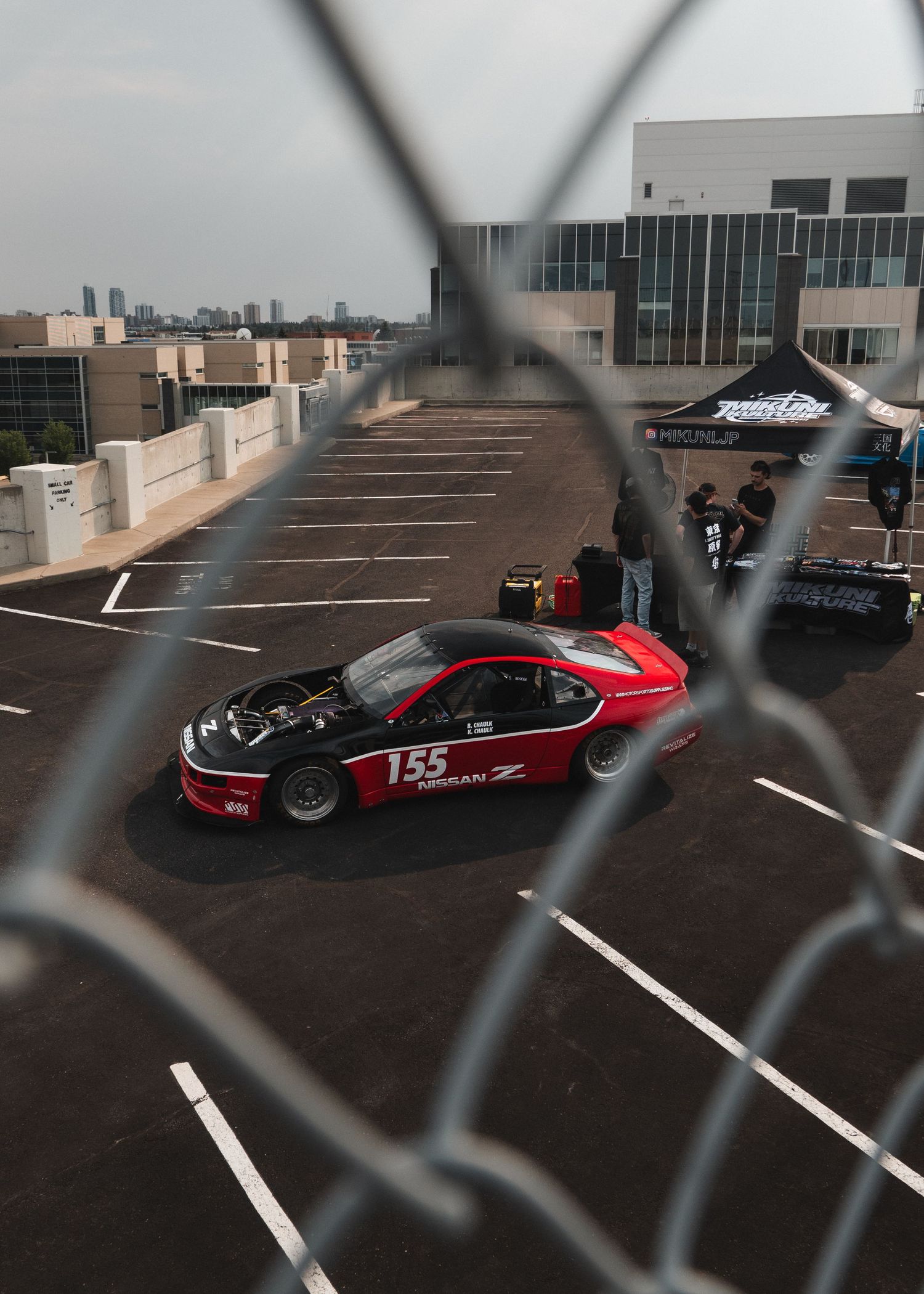 Red Nissan Z race car #155 on rooftop parking lot, viewed through chain-link fence at motorsport event.