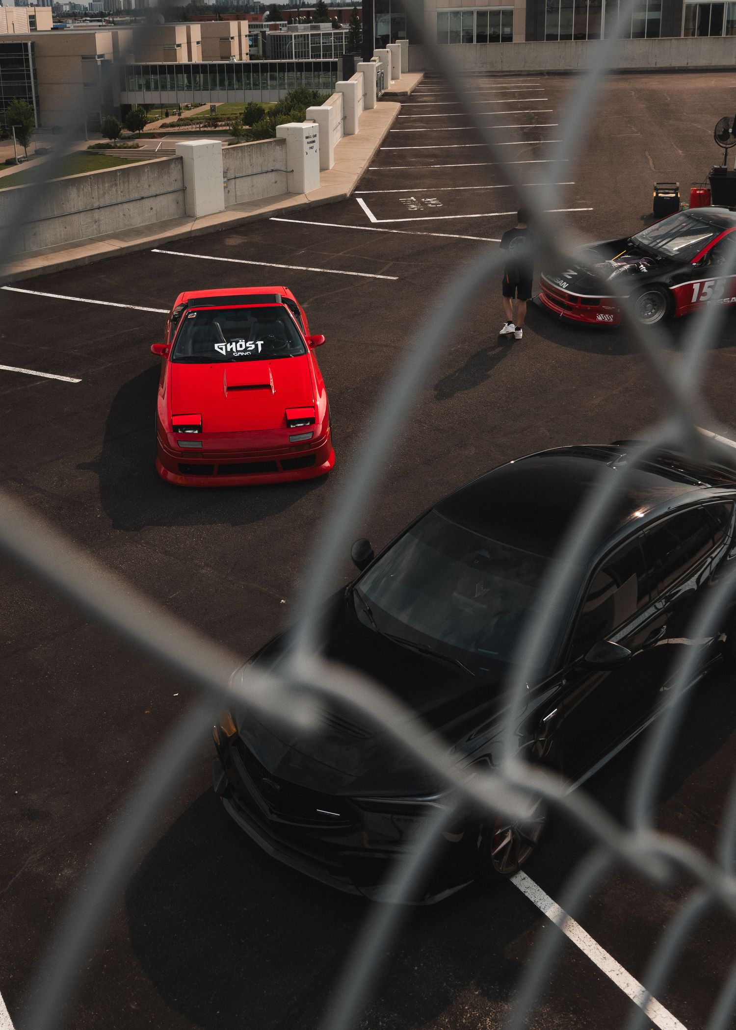Red modified sports car with Ghost decal in parking lot, shot through chain-link fence with black cars nearby.