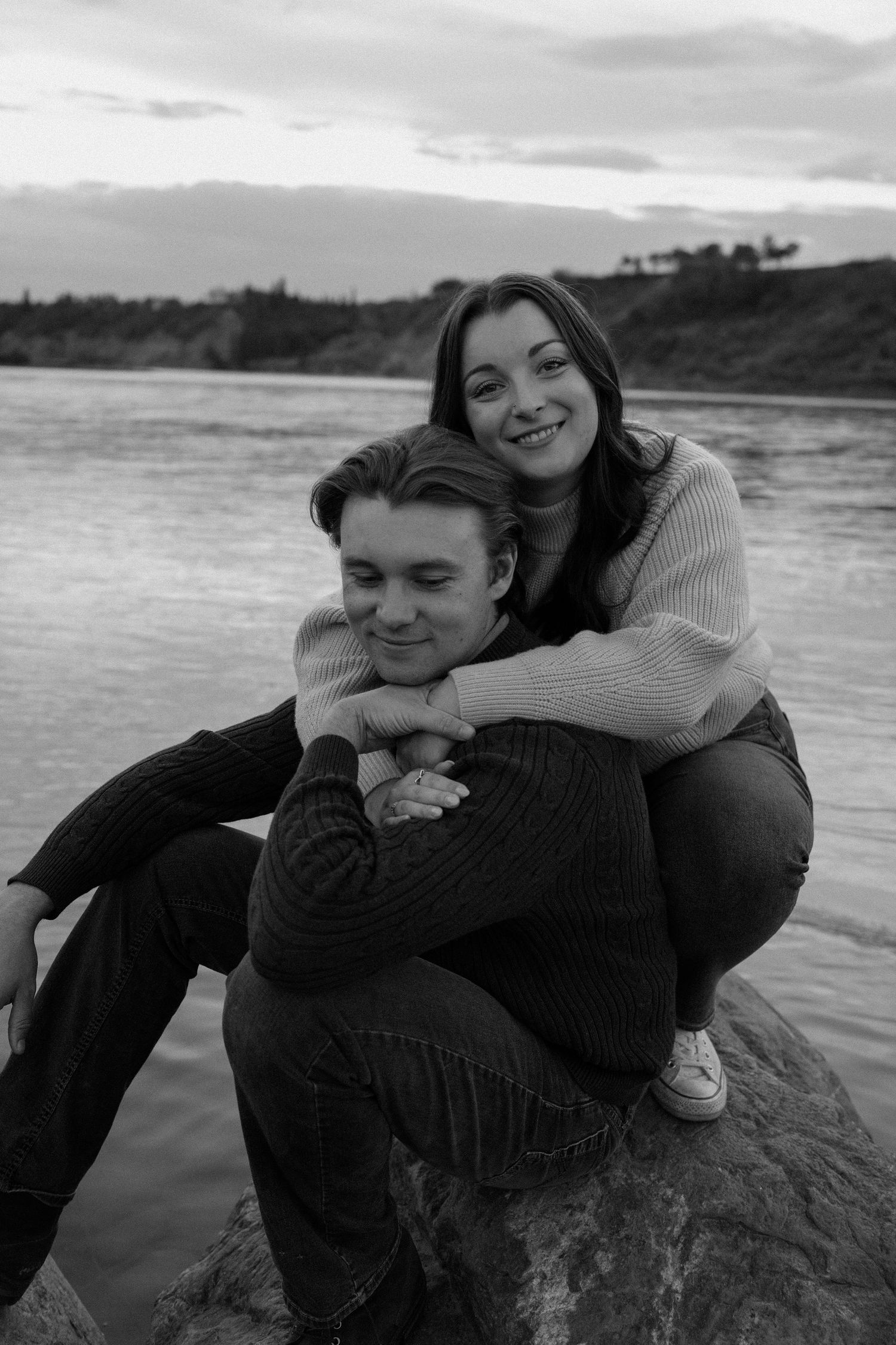 Black and white photo of a happy couple embracing on a rock by a river at sunset.