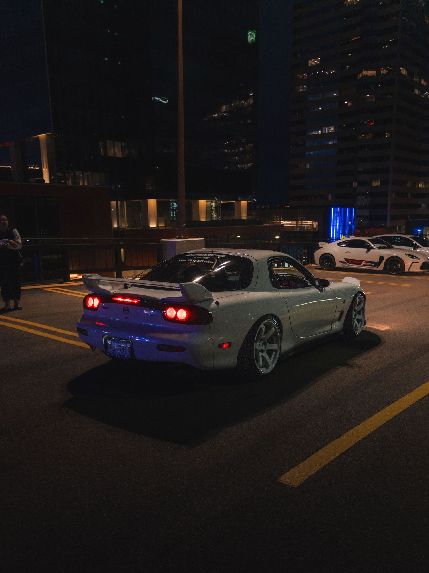 White Mazda RX-7 with glowing red taillights parked on a city street at night with illuminated skyscrapers in the background.