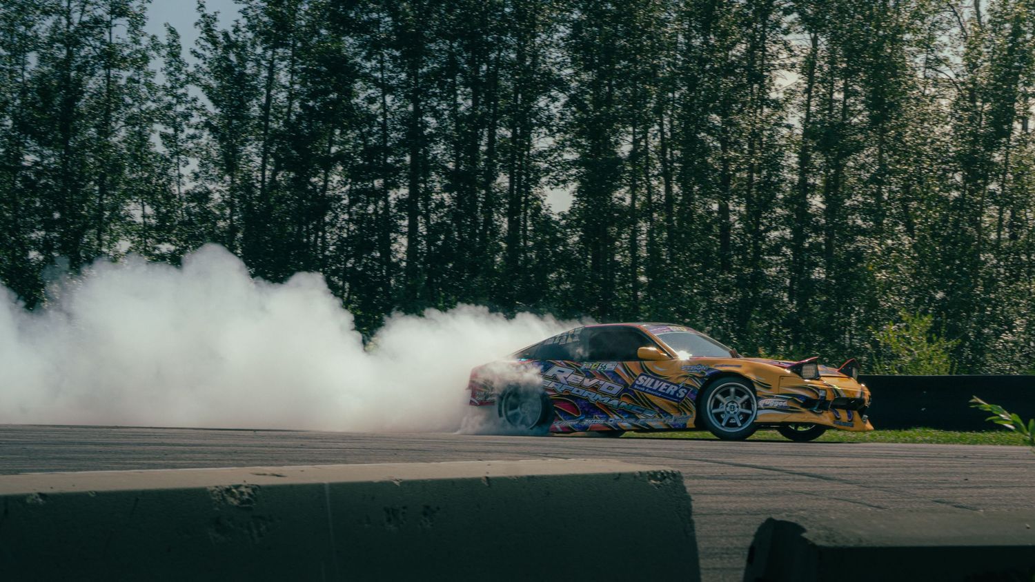 A colorful drift car produces massive smoke clouds while sliding on a race track surrounded by tall green trees.
