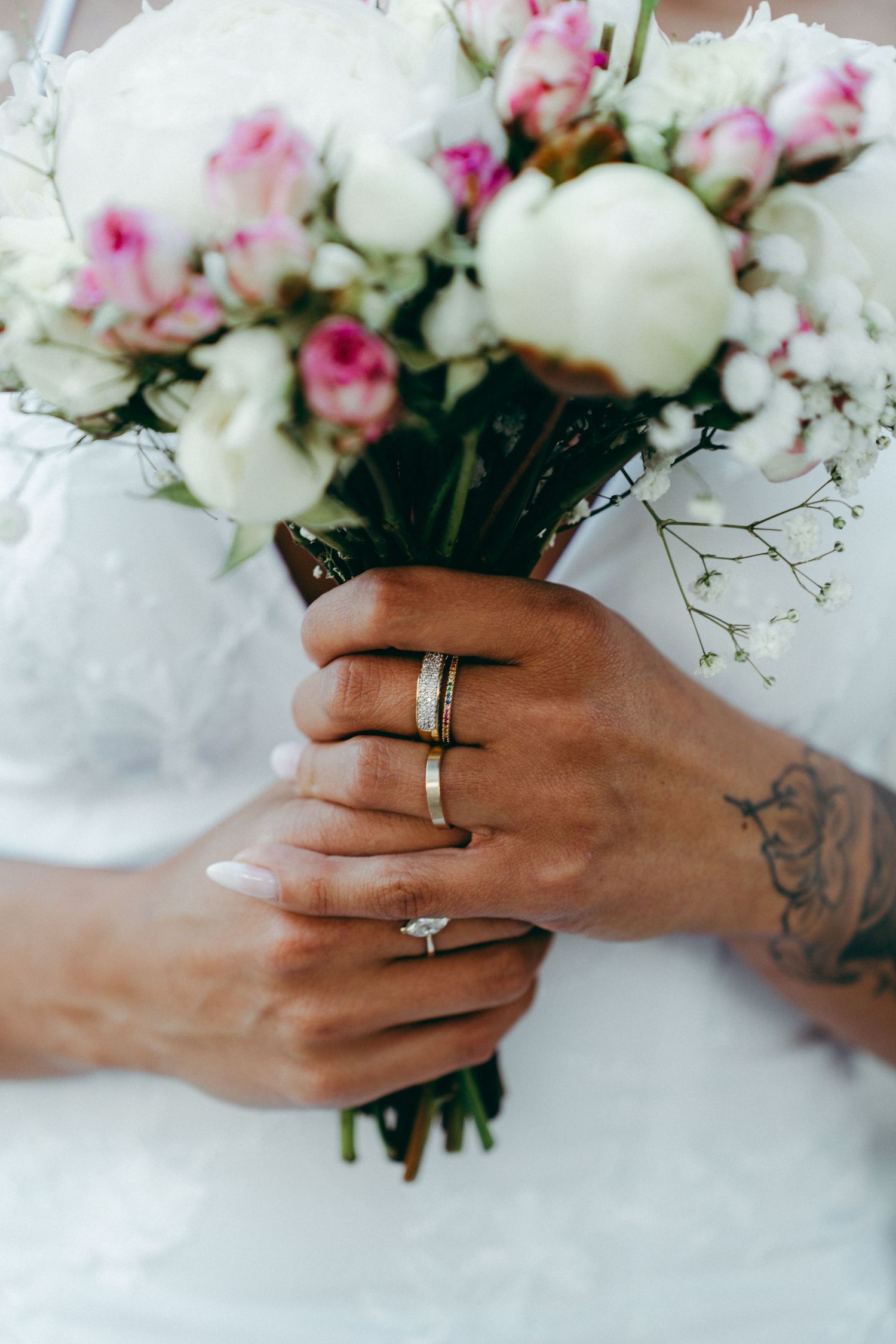 Close-up of tattooed hands holding pink and white wedding bouquet with wedding rings visible.