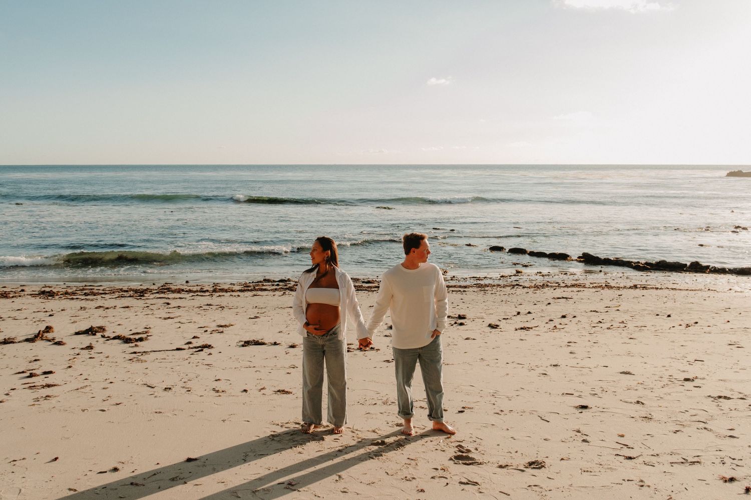 A couple walks along a sandy beach at sunset holding hands with ocean waves in the background.