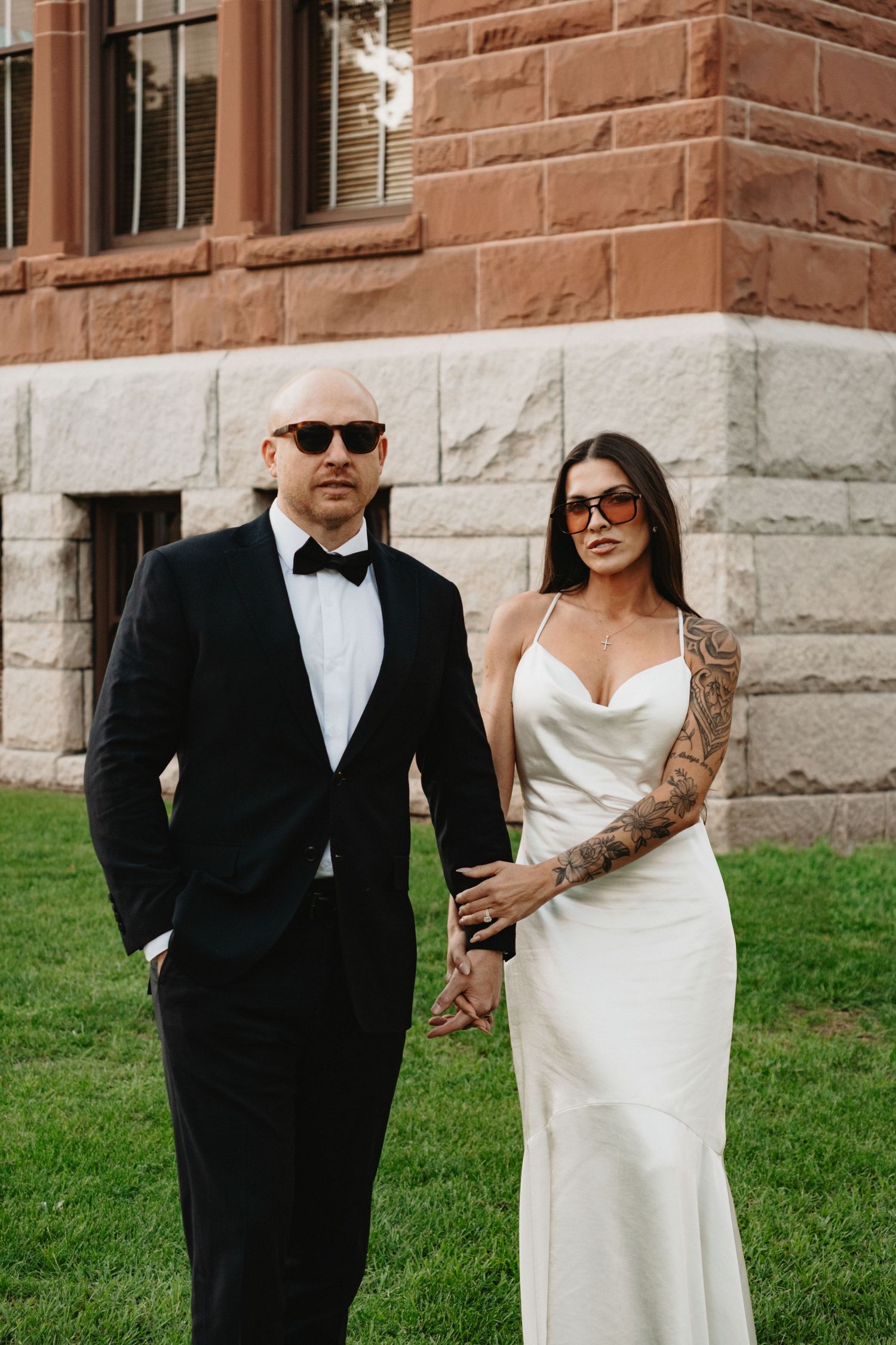 A couple in formal wedding attire walks together on grass in front of a stone building.