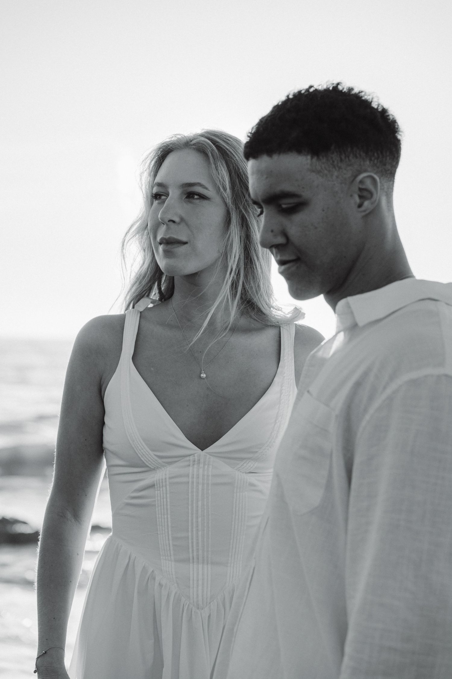A romantic couple in white outfits poses together in a series of intimate black and white beach photos.