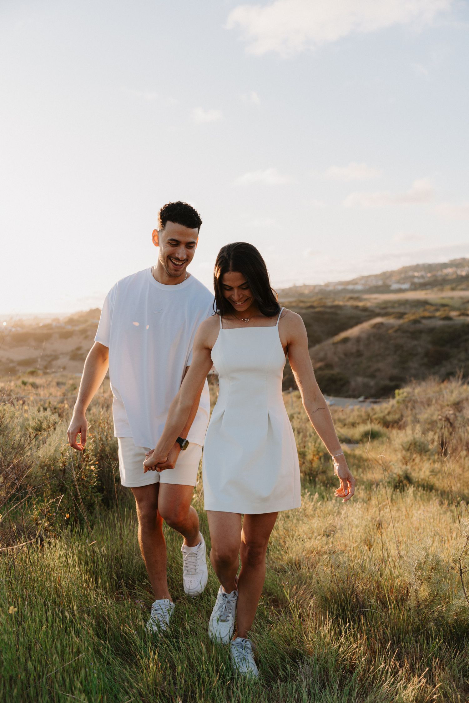 A couple in matching white outfits holding hands while walking through a grassy hillside at sunset.