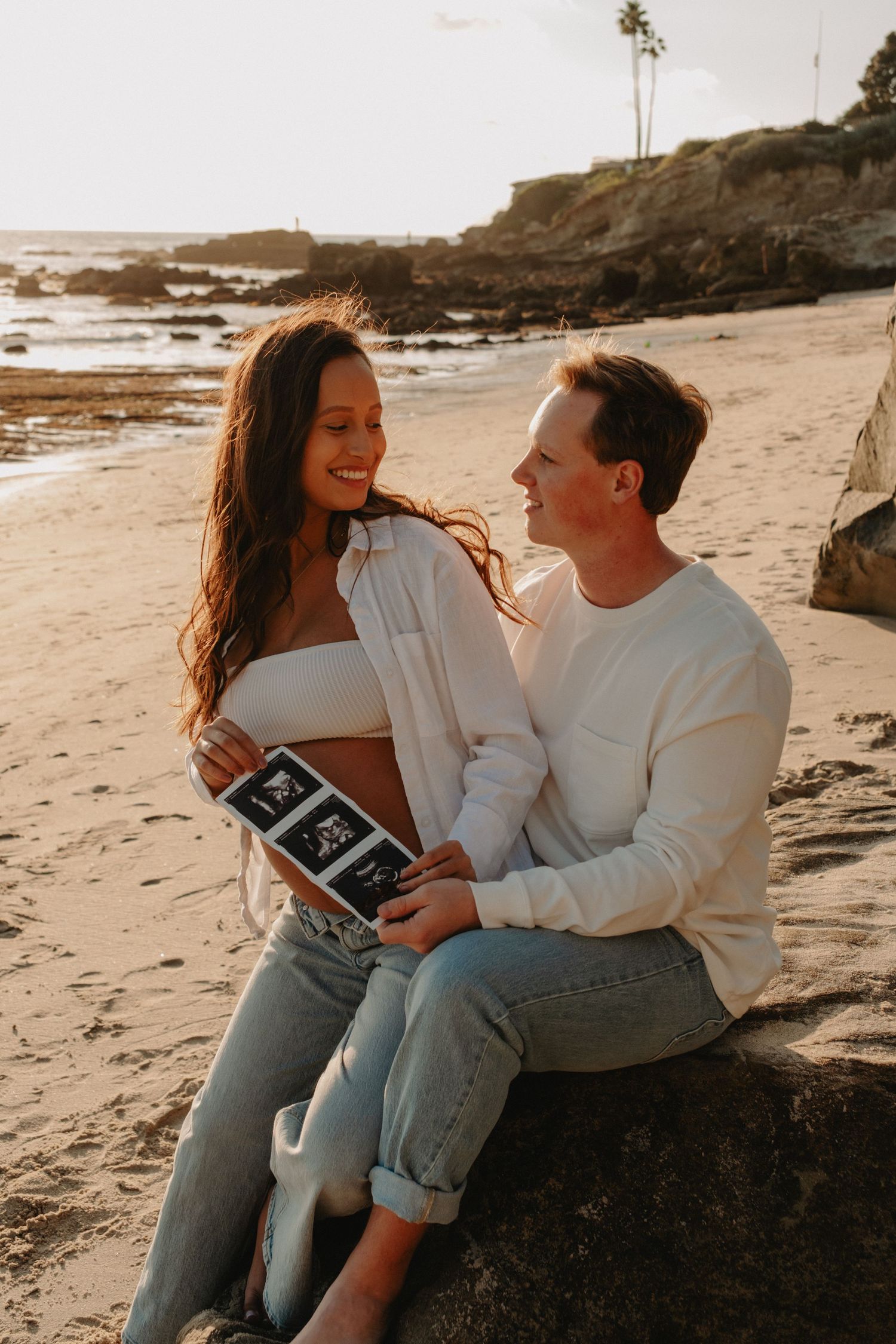 A couple sits on a beach rock at sunset holding ultrasound photos during a pregnancy announcement photoshoot.