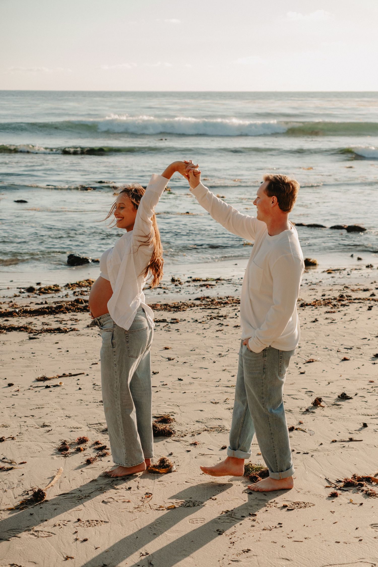 Two people in casual white shirts and jeans dancing together on a beach at sunset with ocean waves in the background.