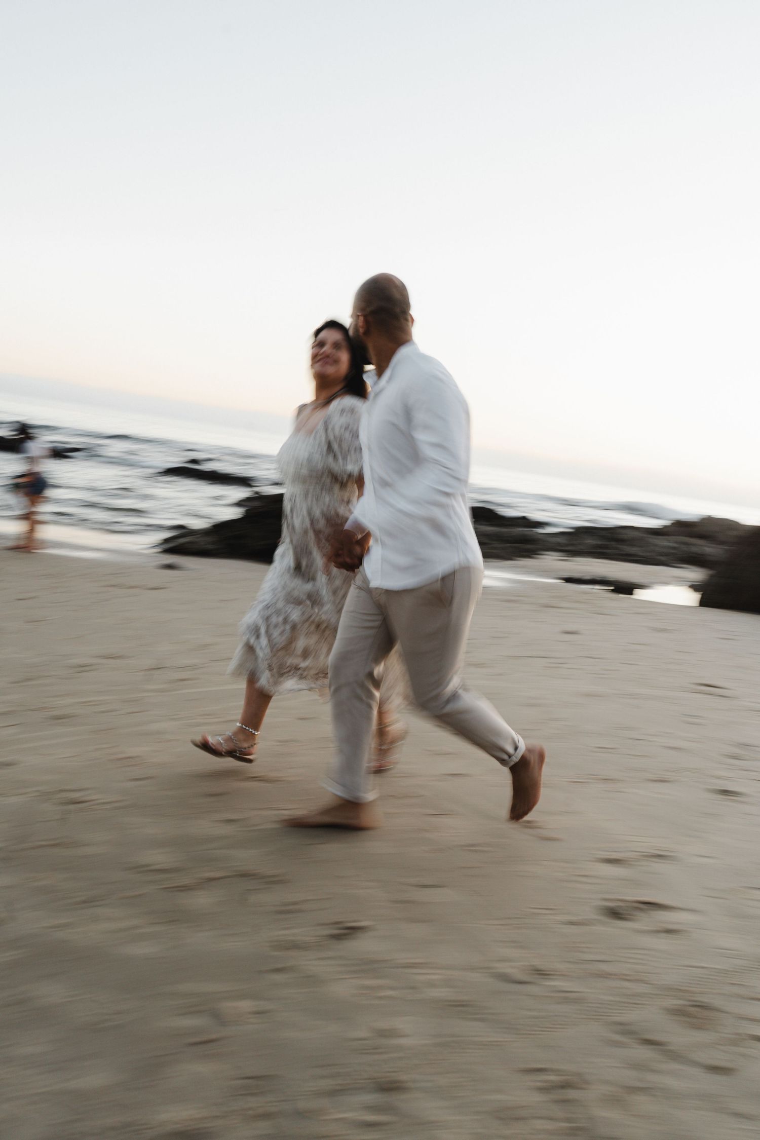 A couple in white clothing dances together on a sandy beach at sunset, creating a romantic silhouette.