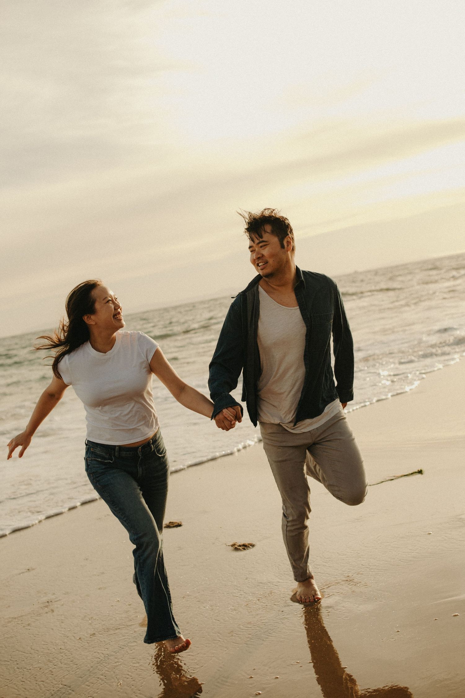 Happy couple holding hands and running barefoot on a sandy beach at sunset with ocean waves in the background.