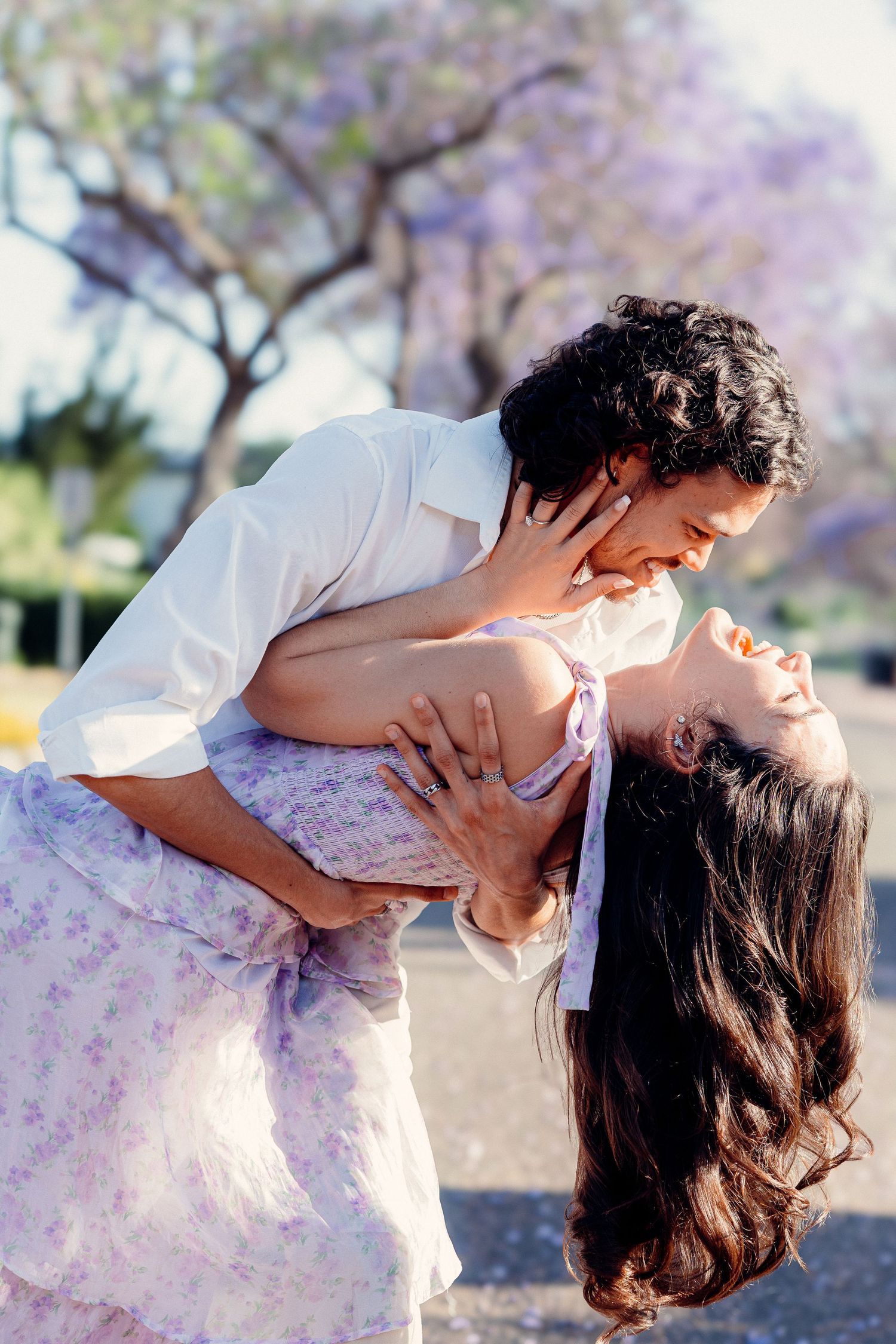Romantic couple poses playfully under flowering jacaranda trees in white outfits.