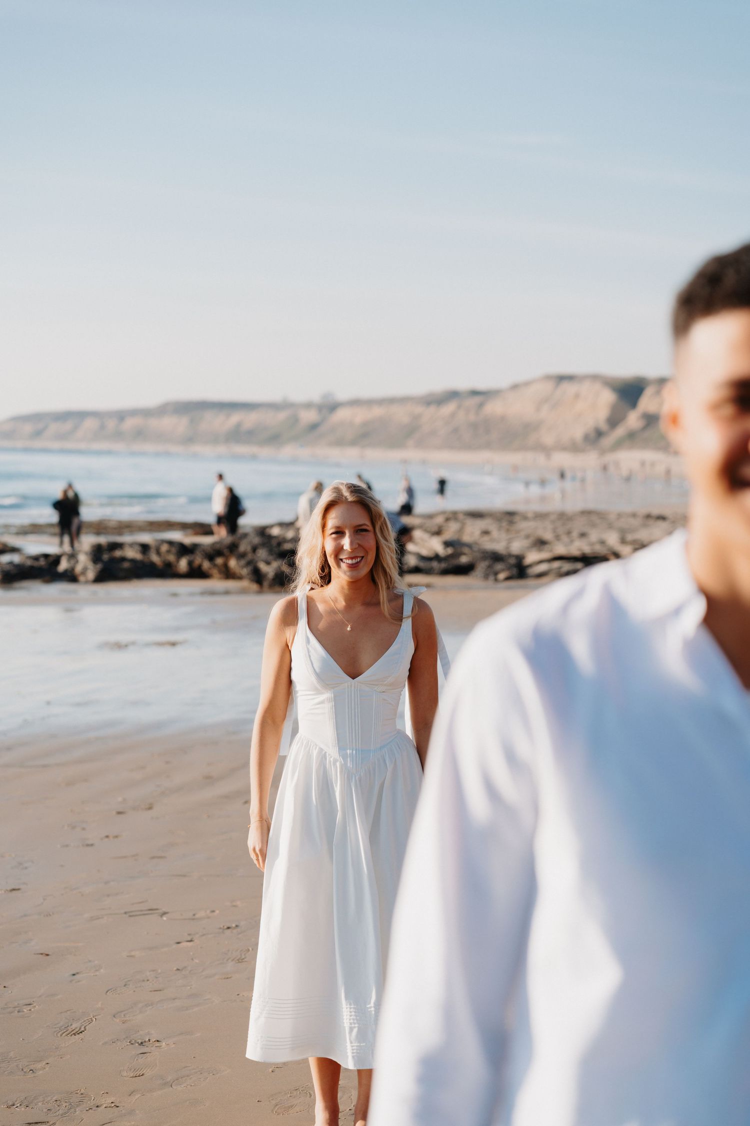 Couple in white outfits walking along scenic beach with coastal cliffs.