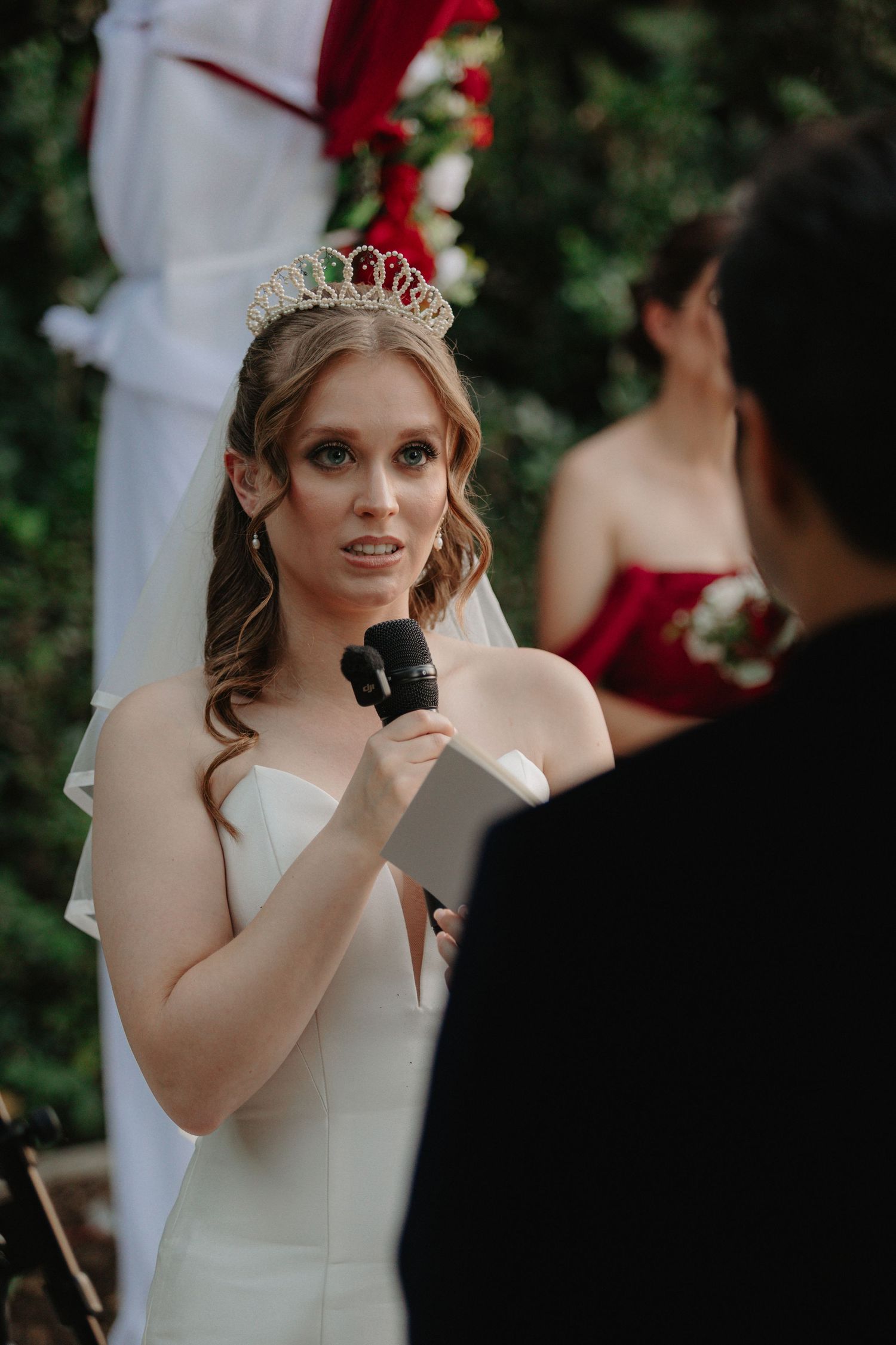 A bride in a white wedding dress and tiara holds a microphone while exchanging vows during an outdoor ceremony.