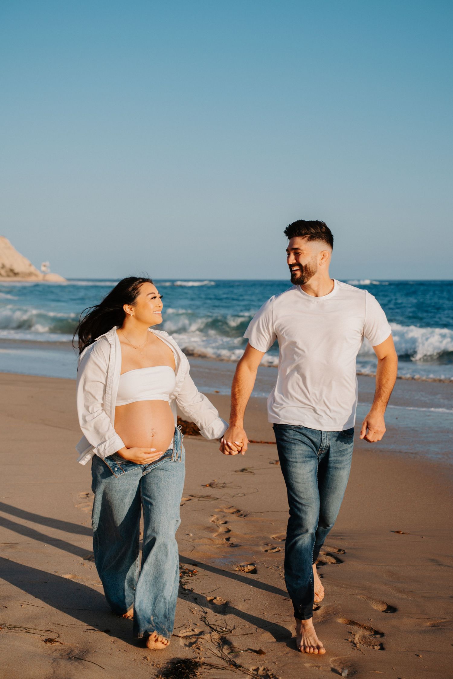 A couple in casual denim and white tops holds hands while walking along the shoreline.