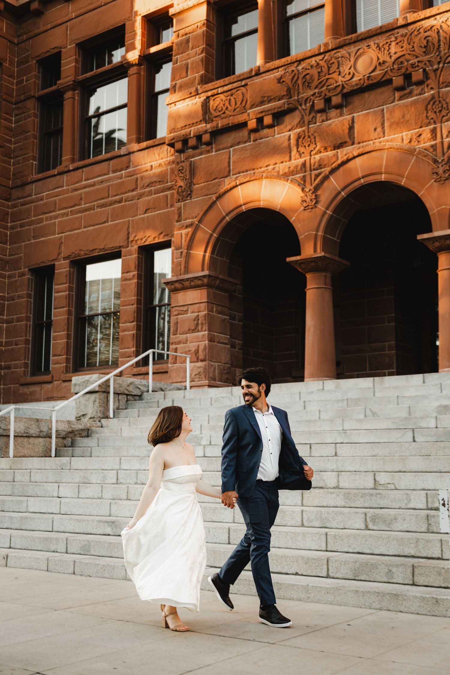 Bride and groom playfully run up stone steps against a red brick building backdrop.