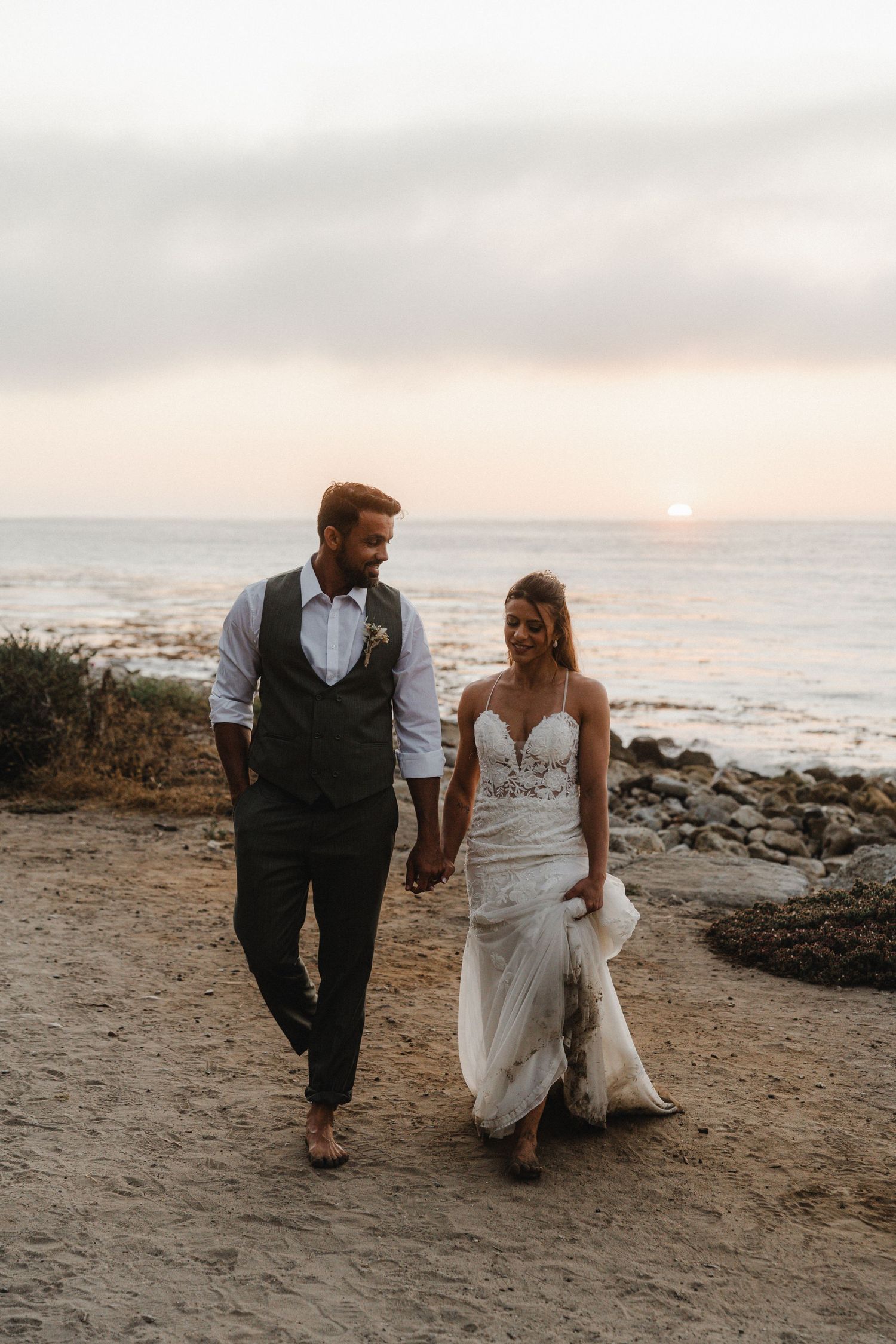 A couple walks hand in hand along a rocky beach at sunset wearing formal wedding attire.