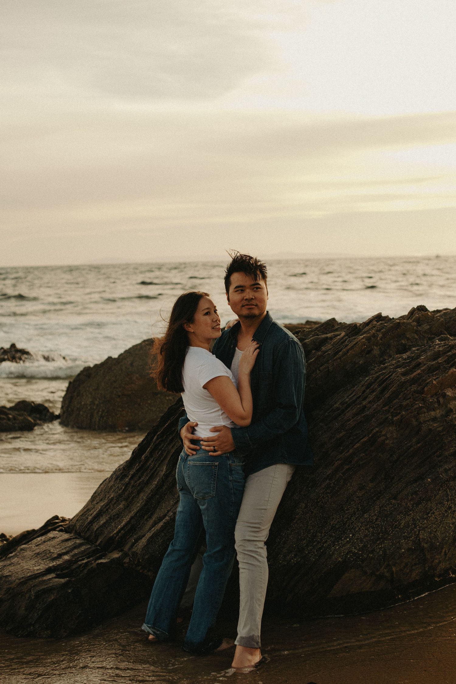 Couple embracing by rocky beach at sunset, woman in white top and jeans, man in dark shirt, ocean waves behind them.