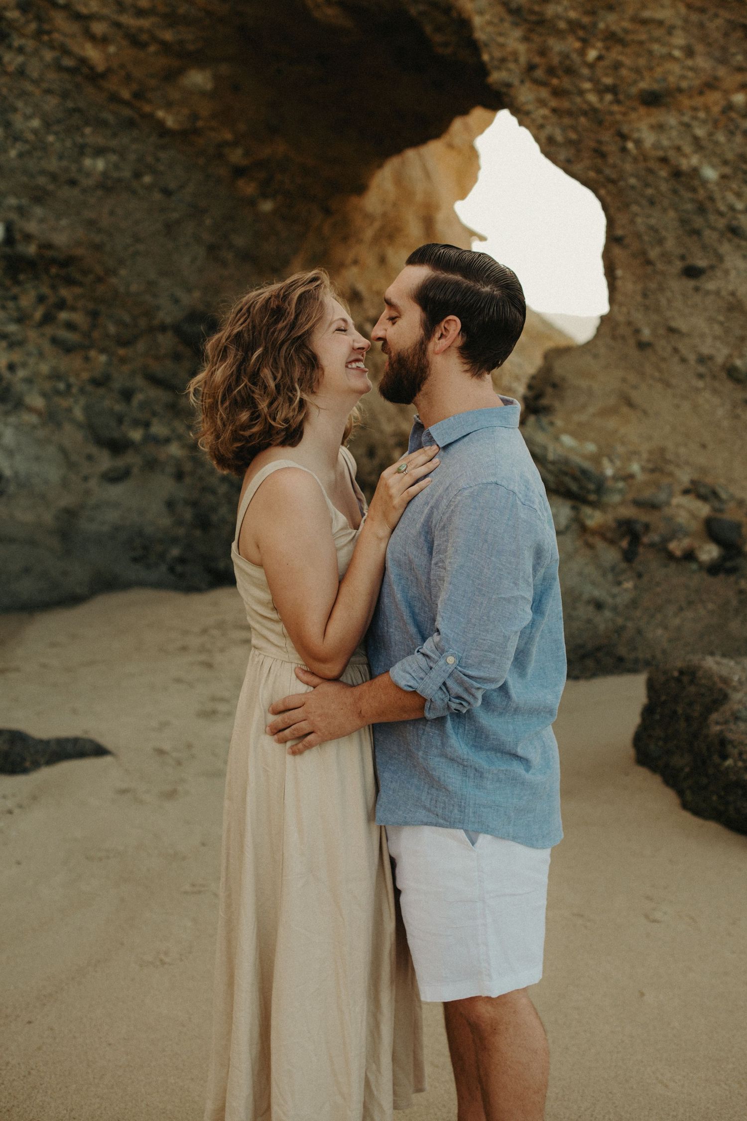 Couple sharing a romantic moment on a sandy beach, embracing near rocky cliffs in casual summer attire.
