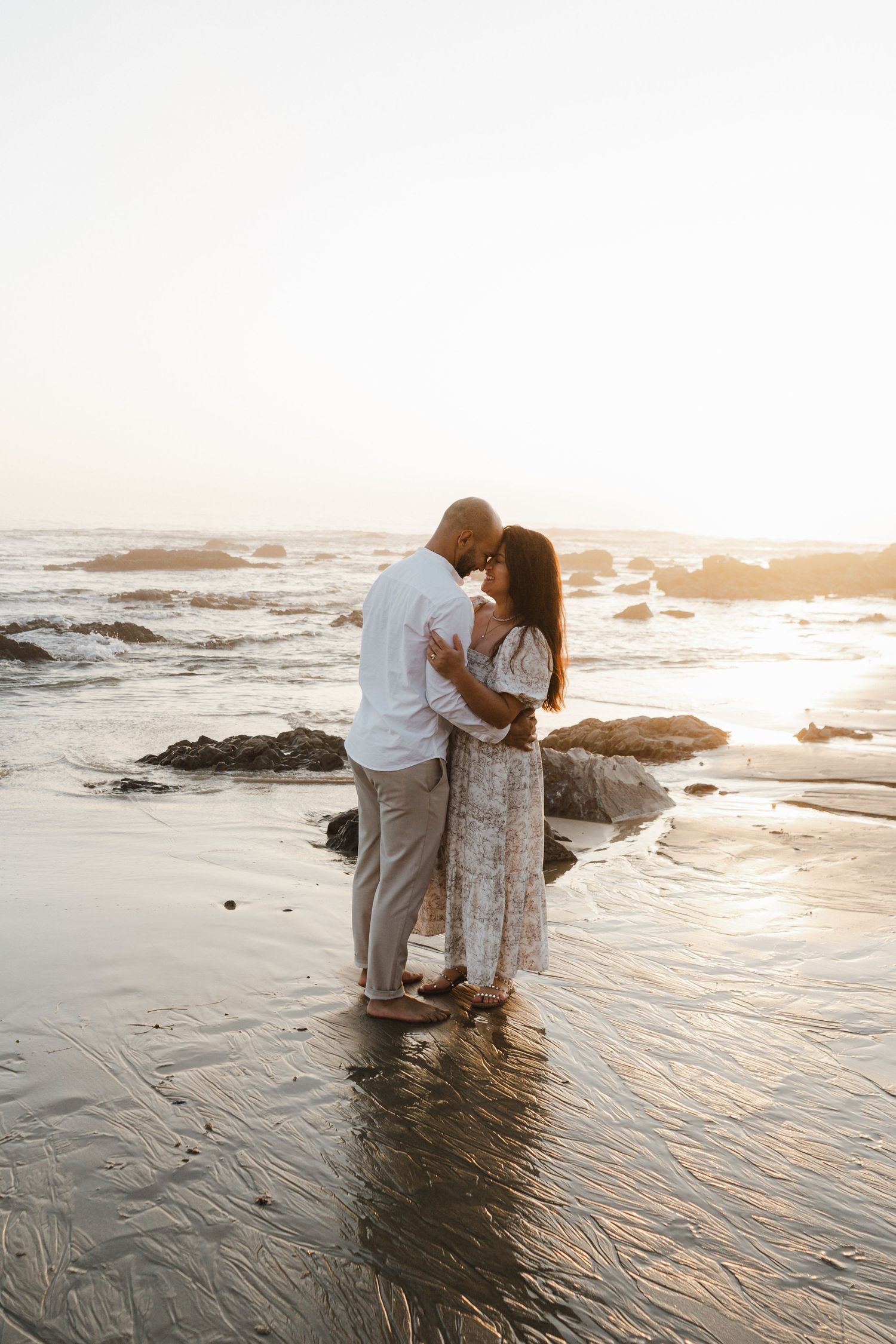 A couple embraces on a sandy beach at sunset as gentle waves wash around their feet.