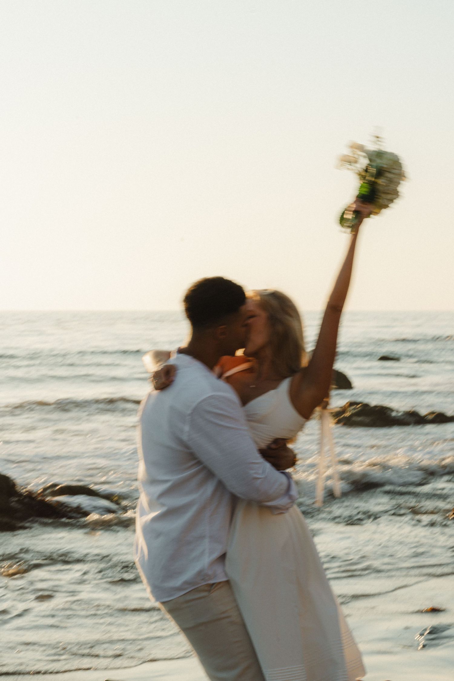 A romantic couple embraces on a rocky beach at sunset while waves crash against the shore in the background.