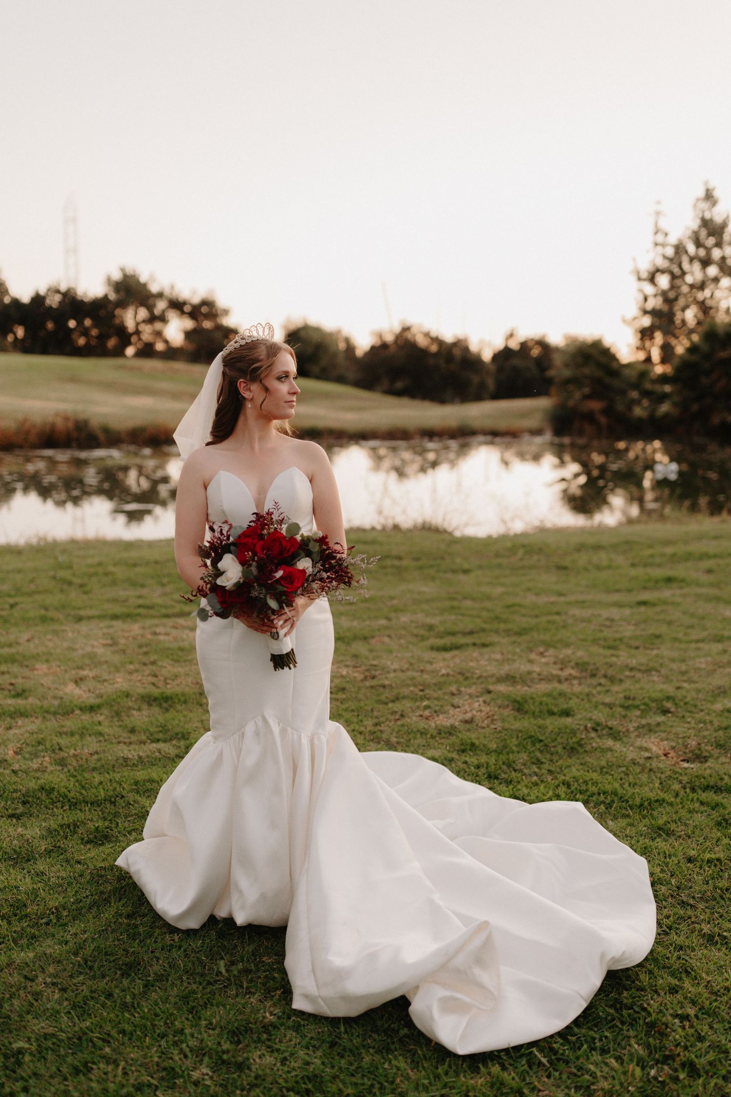 A bride in a flowing white wedding dress poses on a grassy hill at sunset while holding a deep red bouquet.