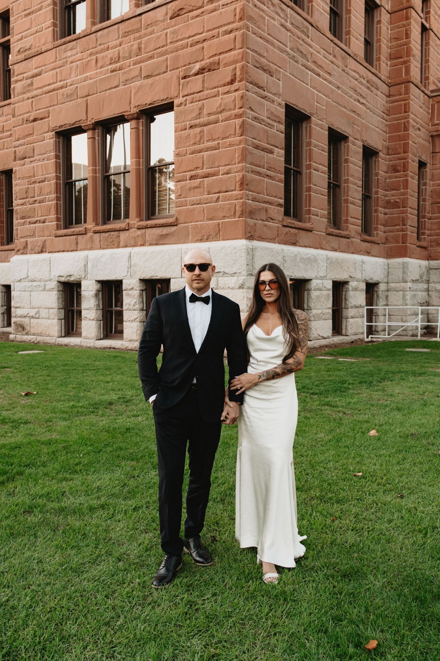 A couple in formal wedding attire walking together on a green lawn next to a brick building exterior.