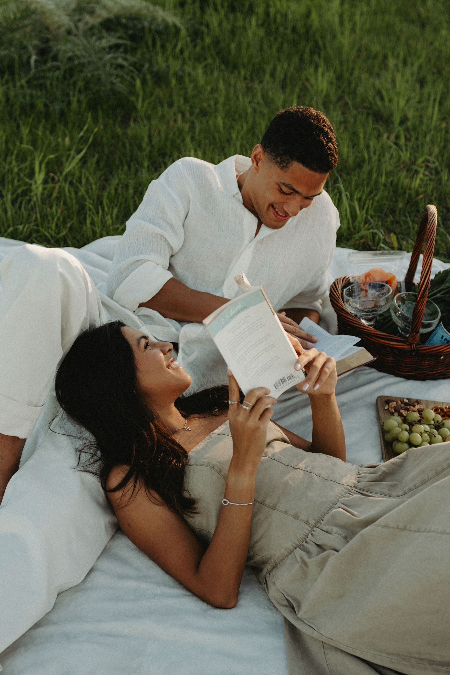 A couple relaxes together on a white blanket while looking at their phones in a grassy outdoor setting.
