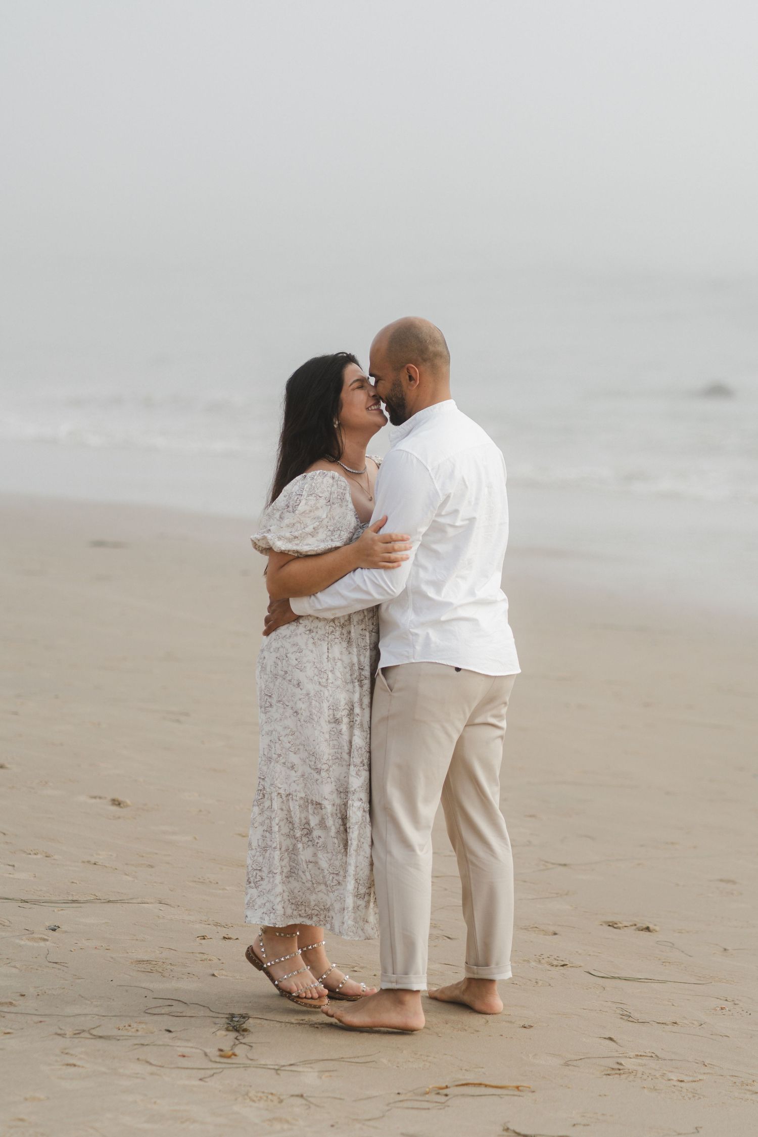 Couple shares romantic moment on sandy beach in light colored attire.