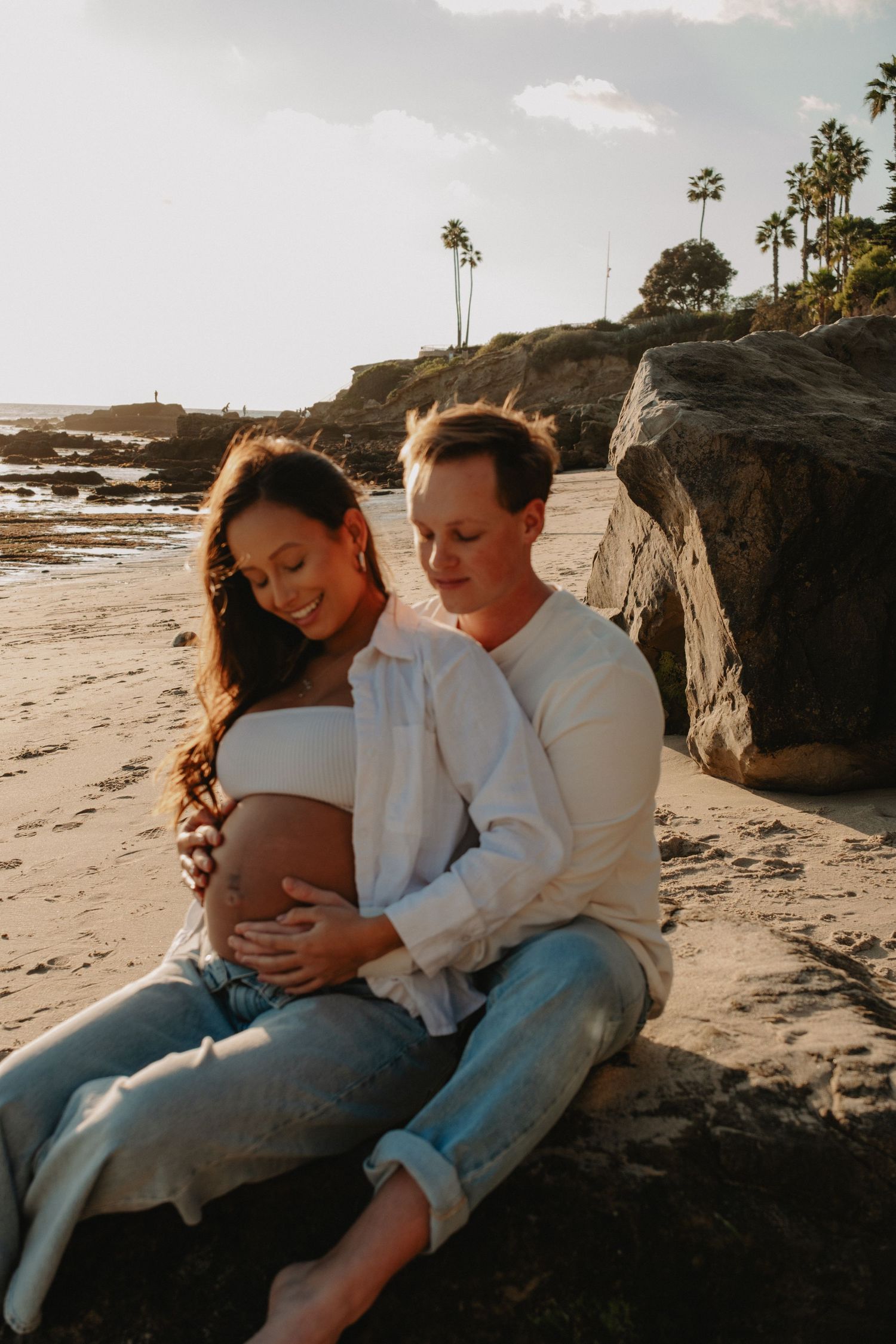 Romantic couple sharing intimate moments on a rocky beach at sunset in matching white outfits.