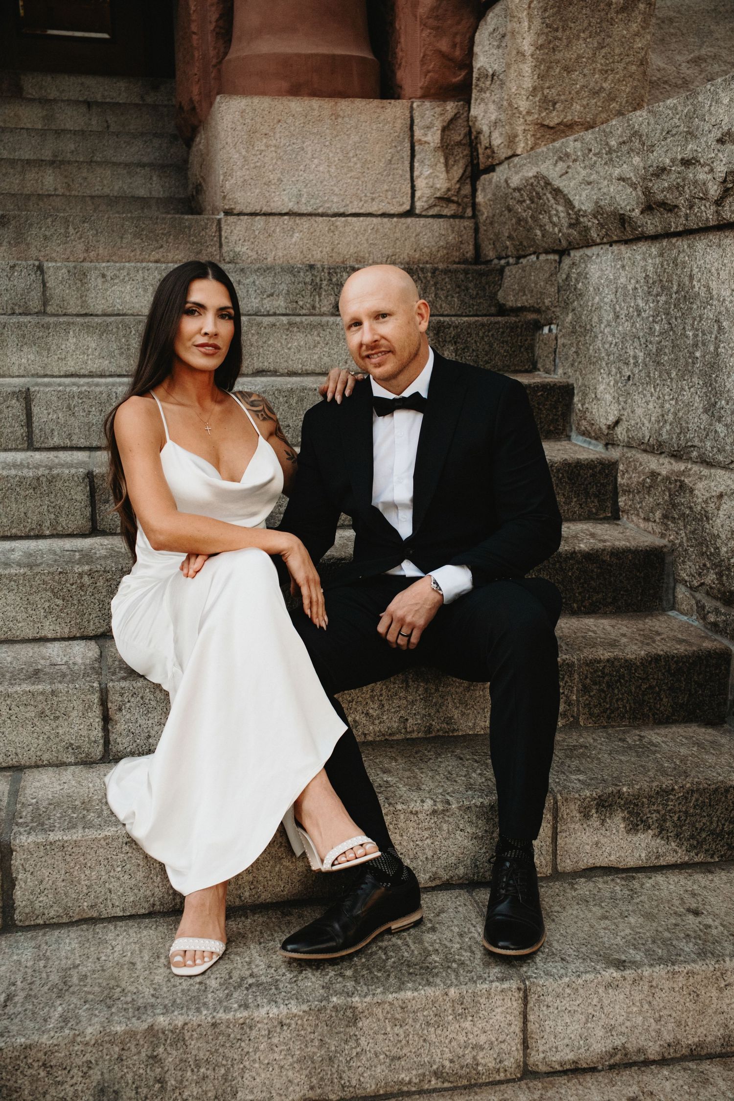 A couple sits intimately on stone steps, with the bride in a white dress and groom in black tuxedo.