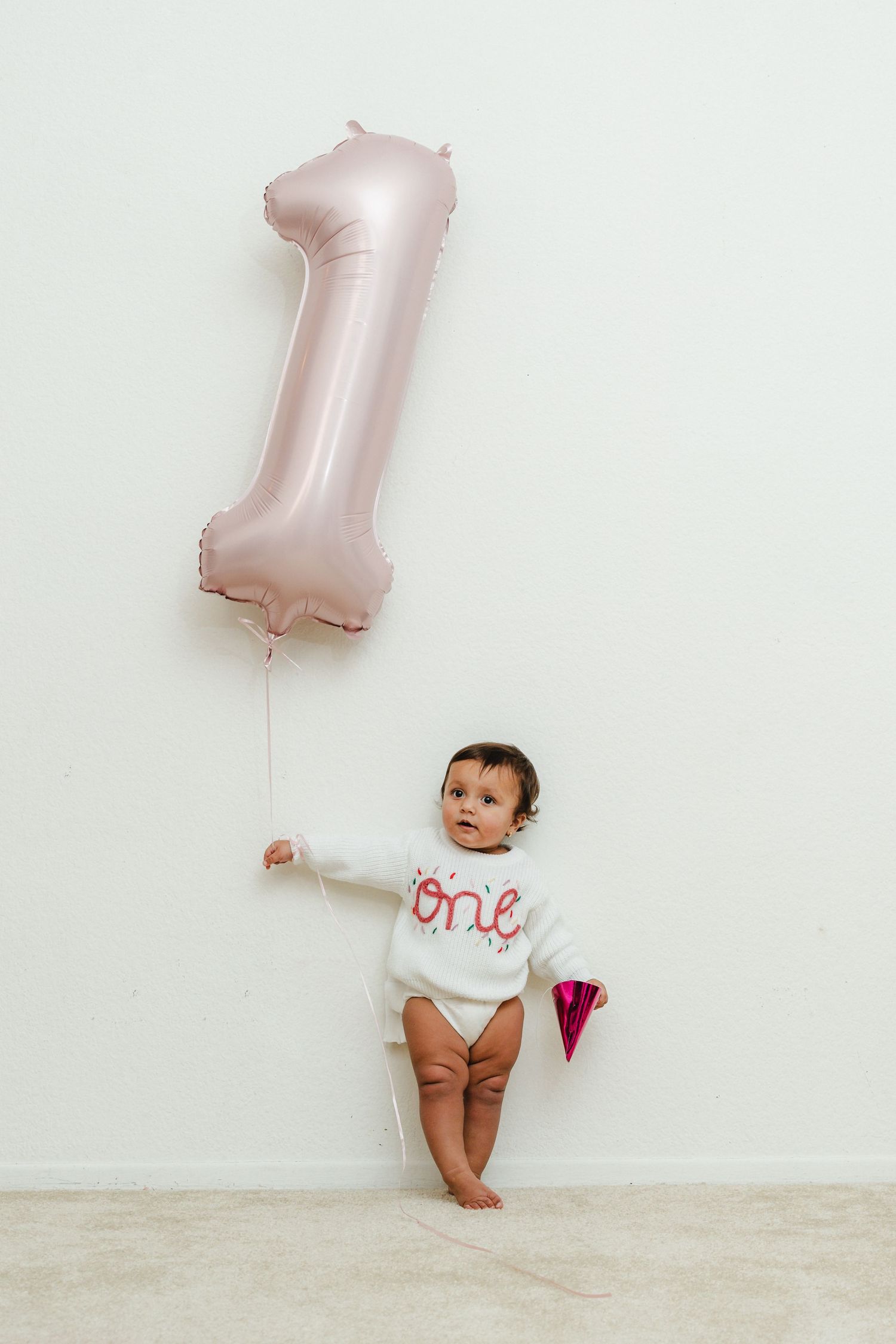 Baby in white outfit poses with pink number one balloon against white wall.