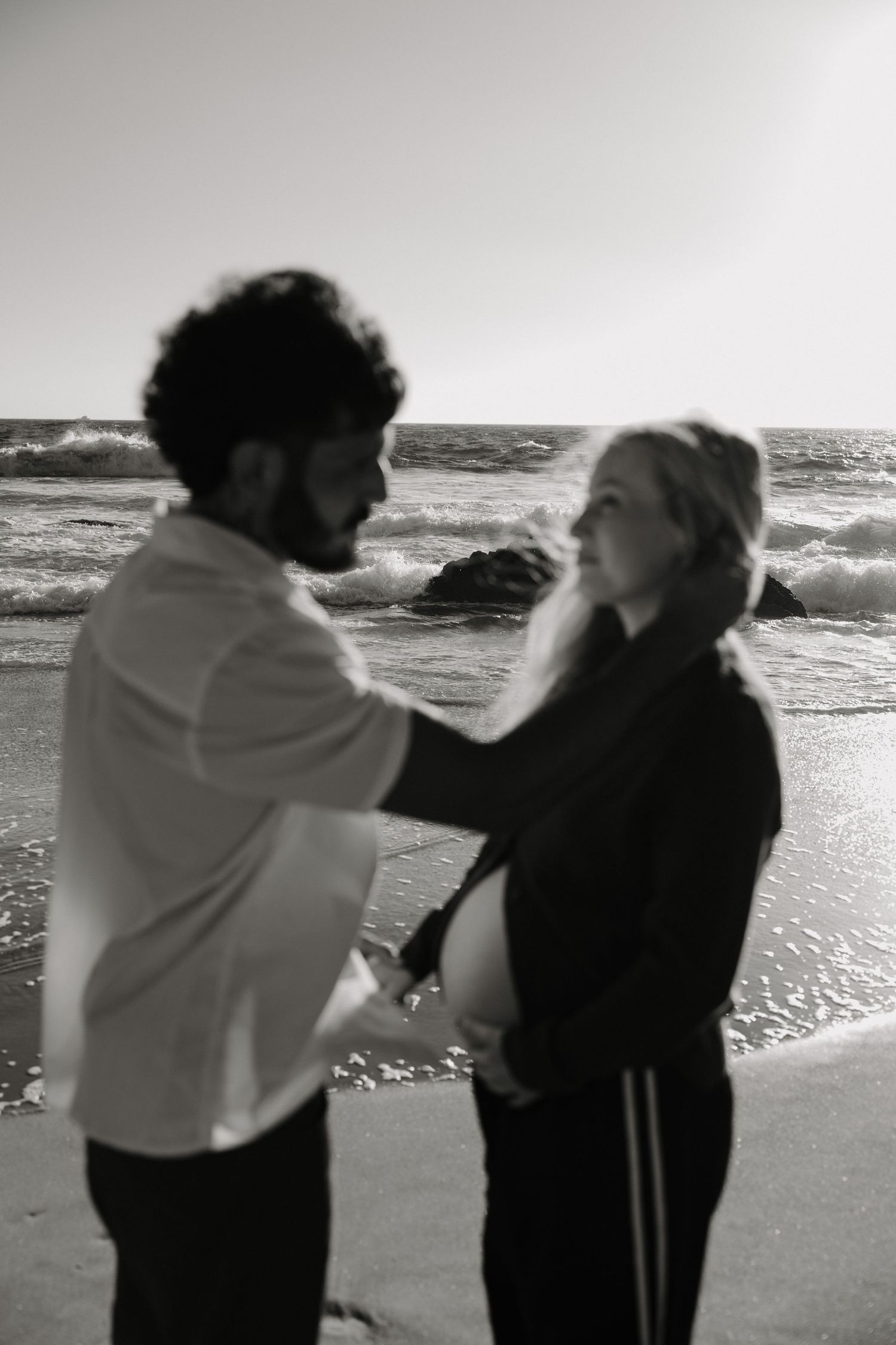 A couple in silhouette playfully interacts on a beach with waves and ocean in the background.